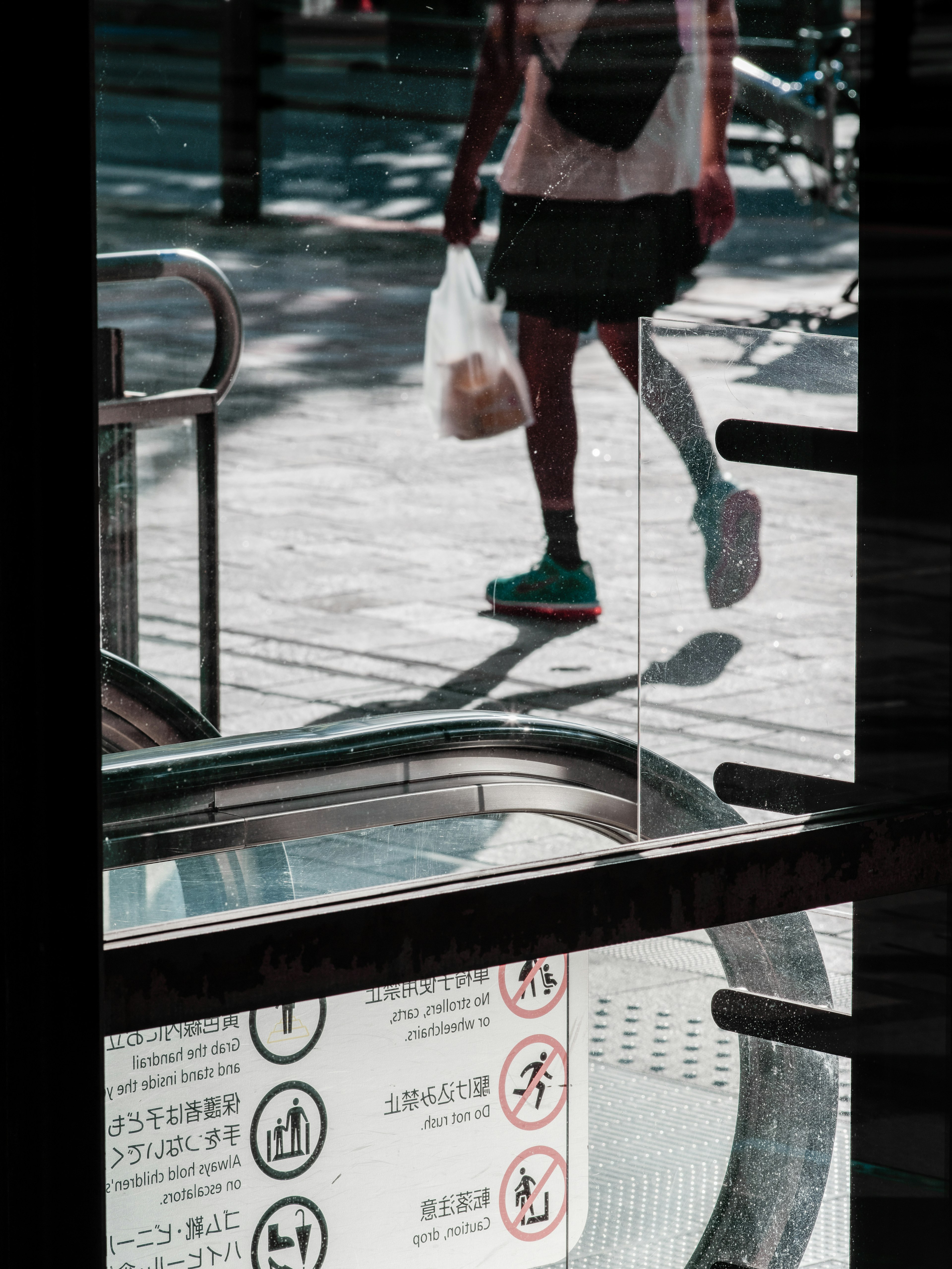 Una persona caminando frente a una escalera mecánica con una bolsa y reflejos de la calle visibles