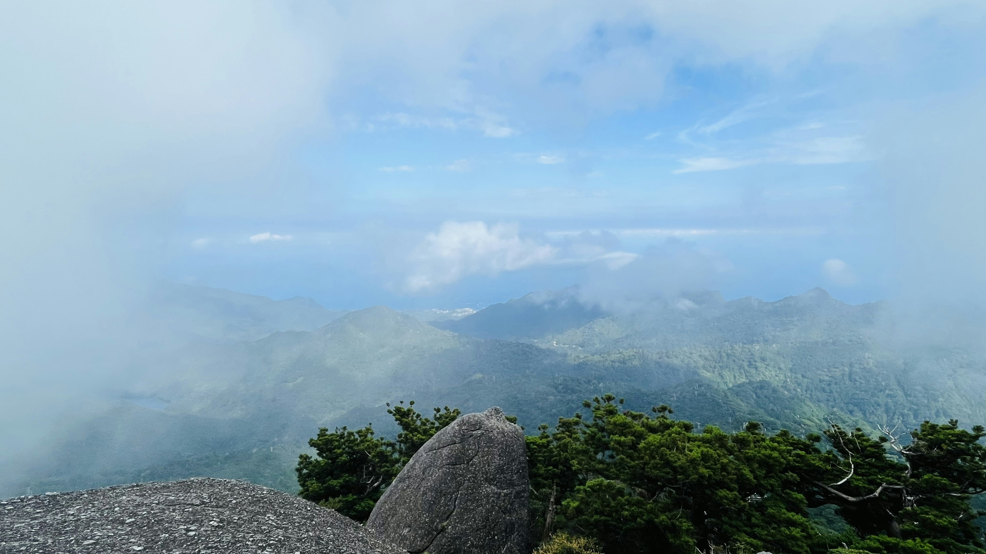 Aussicht von einem Berggipfel mit Wolken blauem Himmel grünen Bäumen und fernen Bergen