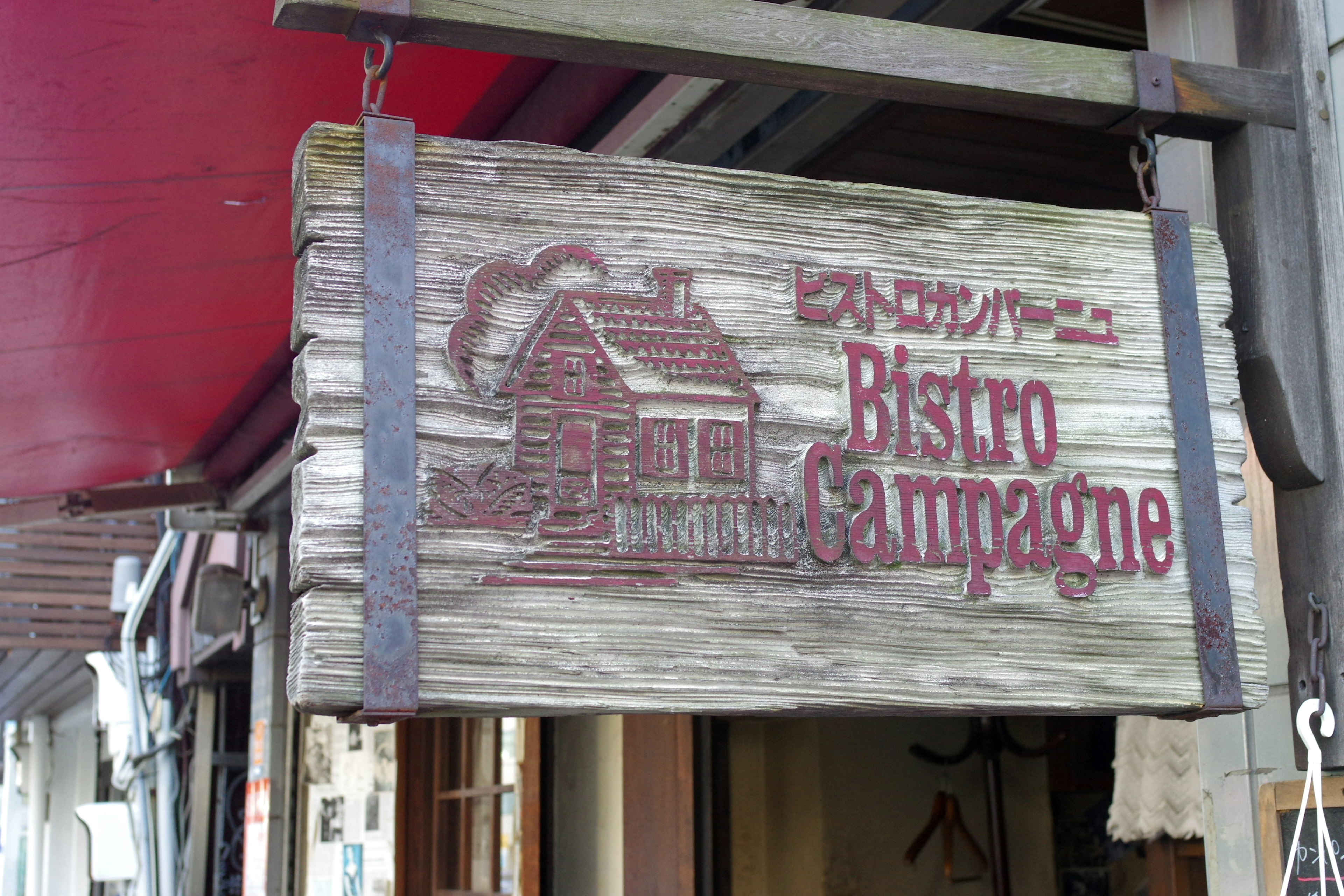 ป้ายไม้สำหรับ Bistro Campagne ที่มีภาพประกอบของบ้านหลังคาแดง