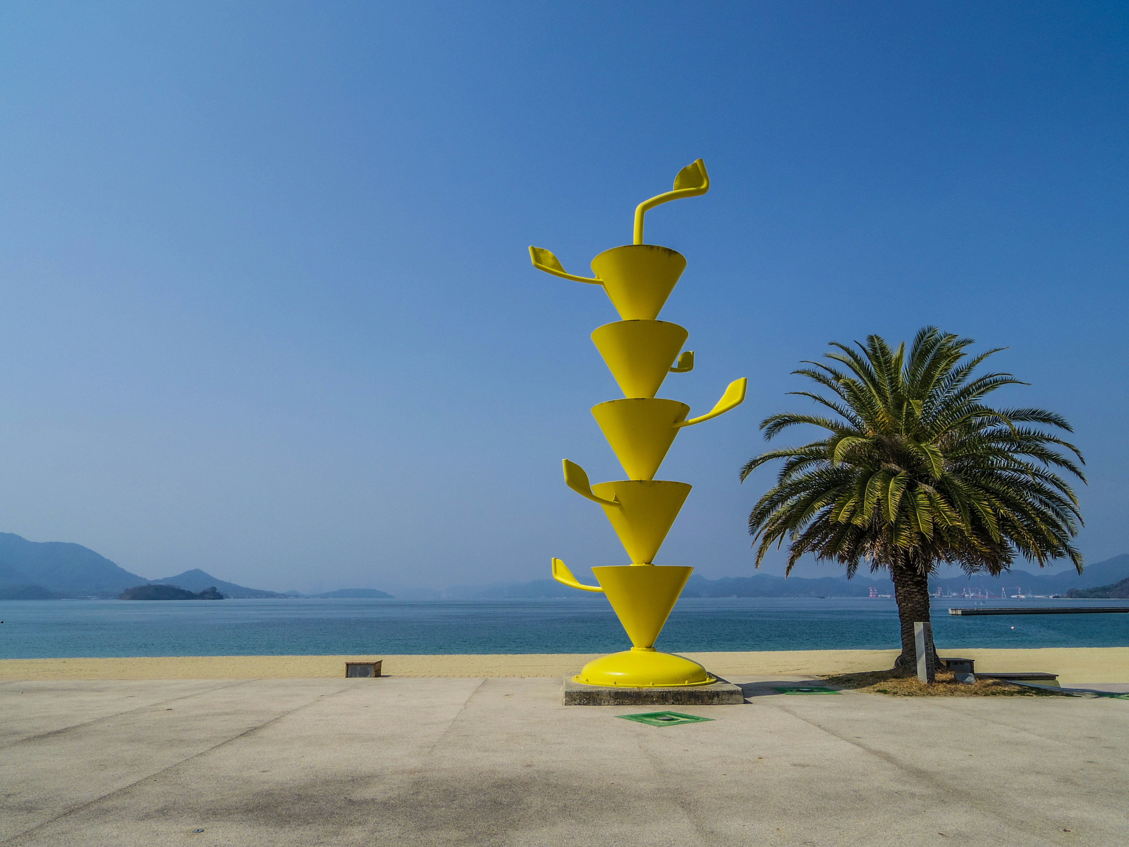 Yellow sculpture with unique shapes beside a palm tree against a blue sky and sea