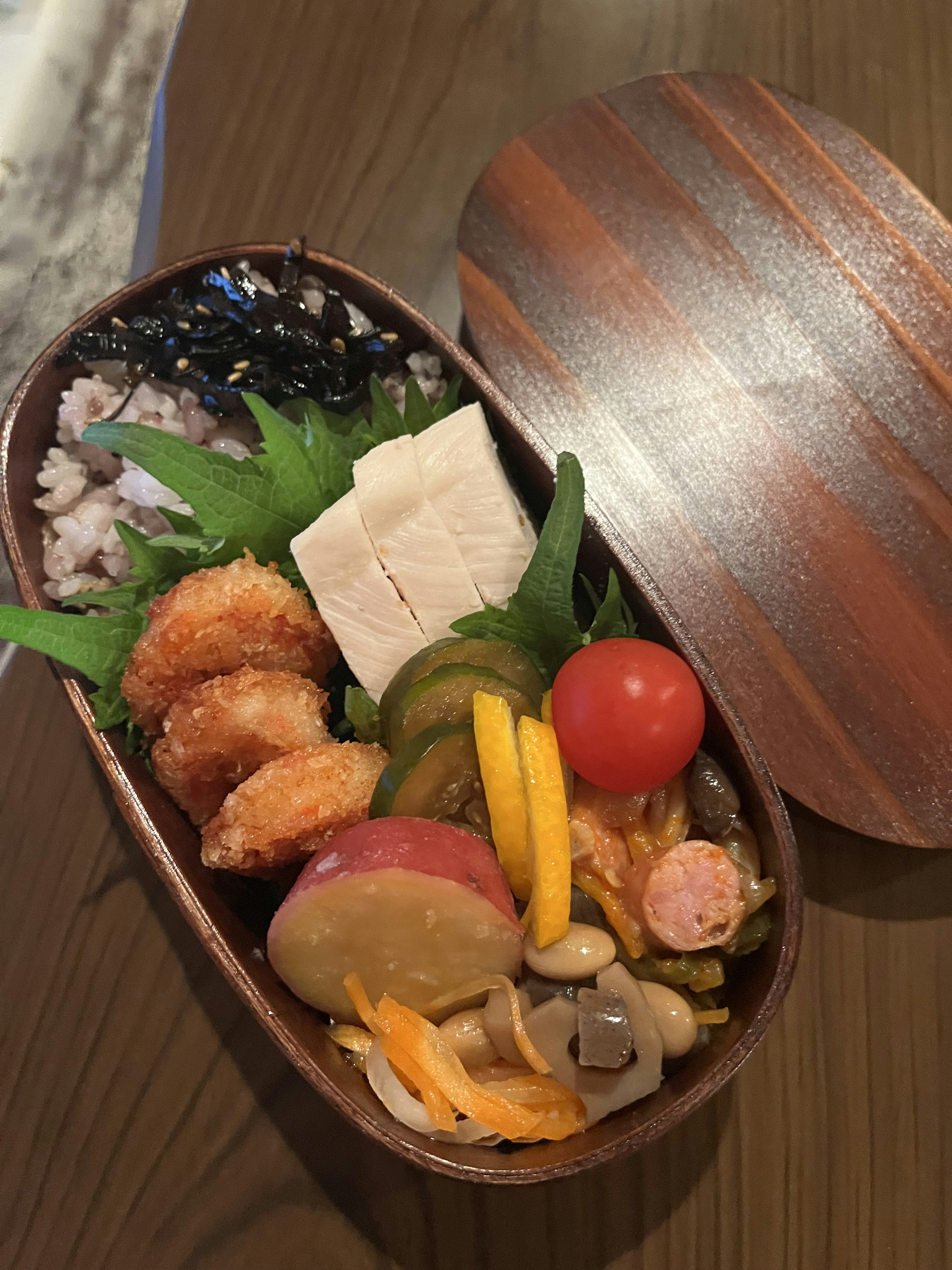 A beautifully arranged bento box featuring colorful dishes including fried shrimp, tofu, vegetables, cherry tomatoes, and fruits