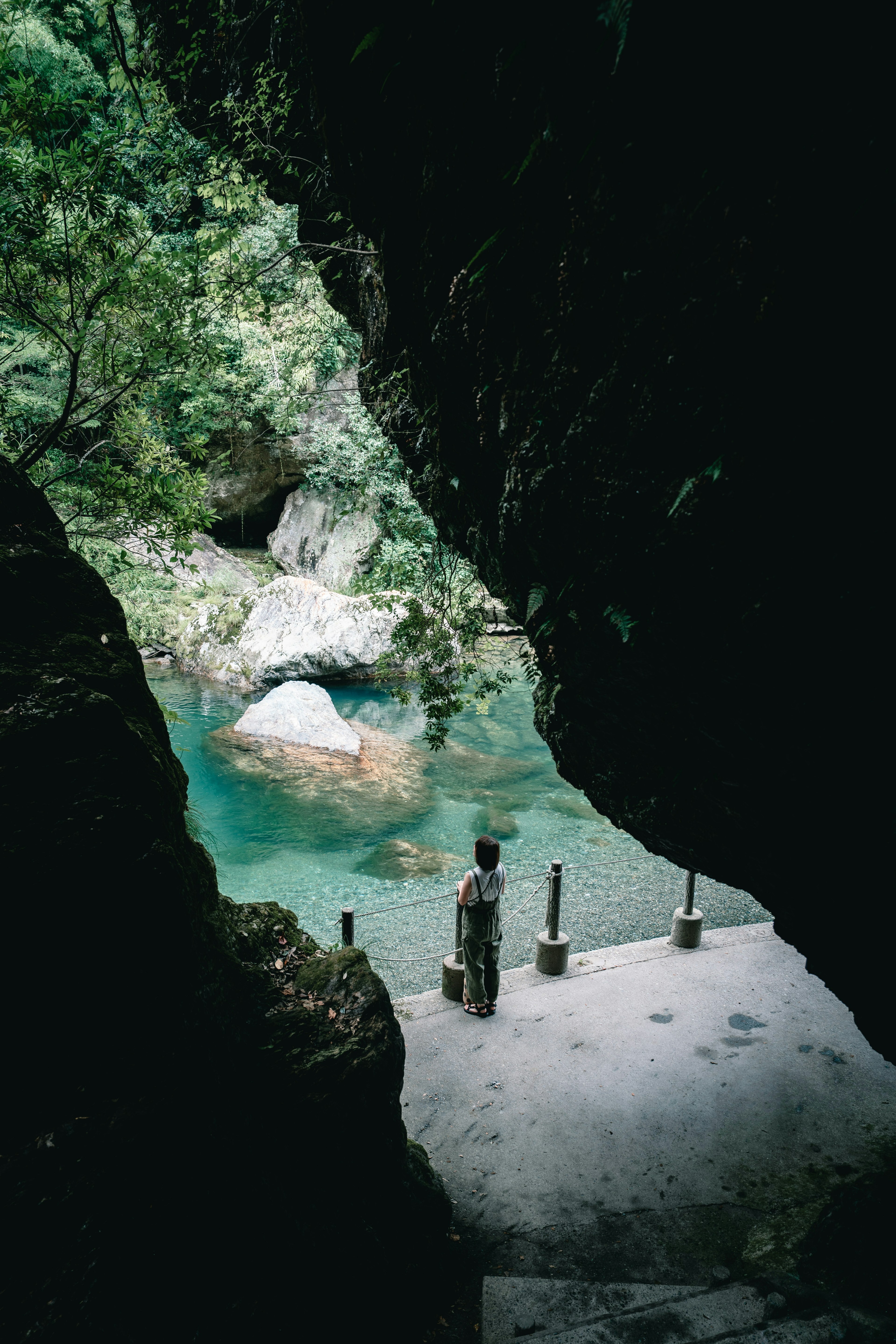 Một người đứng ở cửa hang nhìn ra hồ nước màu ngọc bích yên tĩnh được bao quanh bởi cây cối