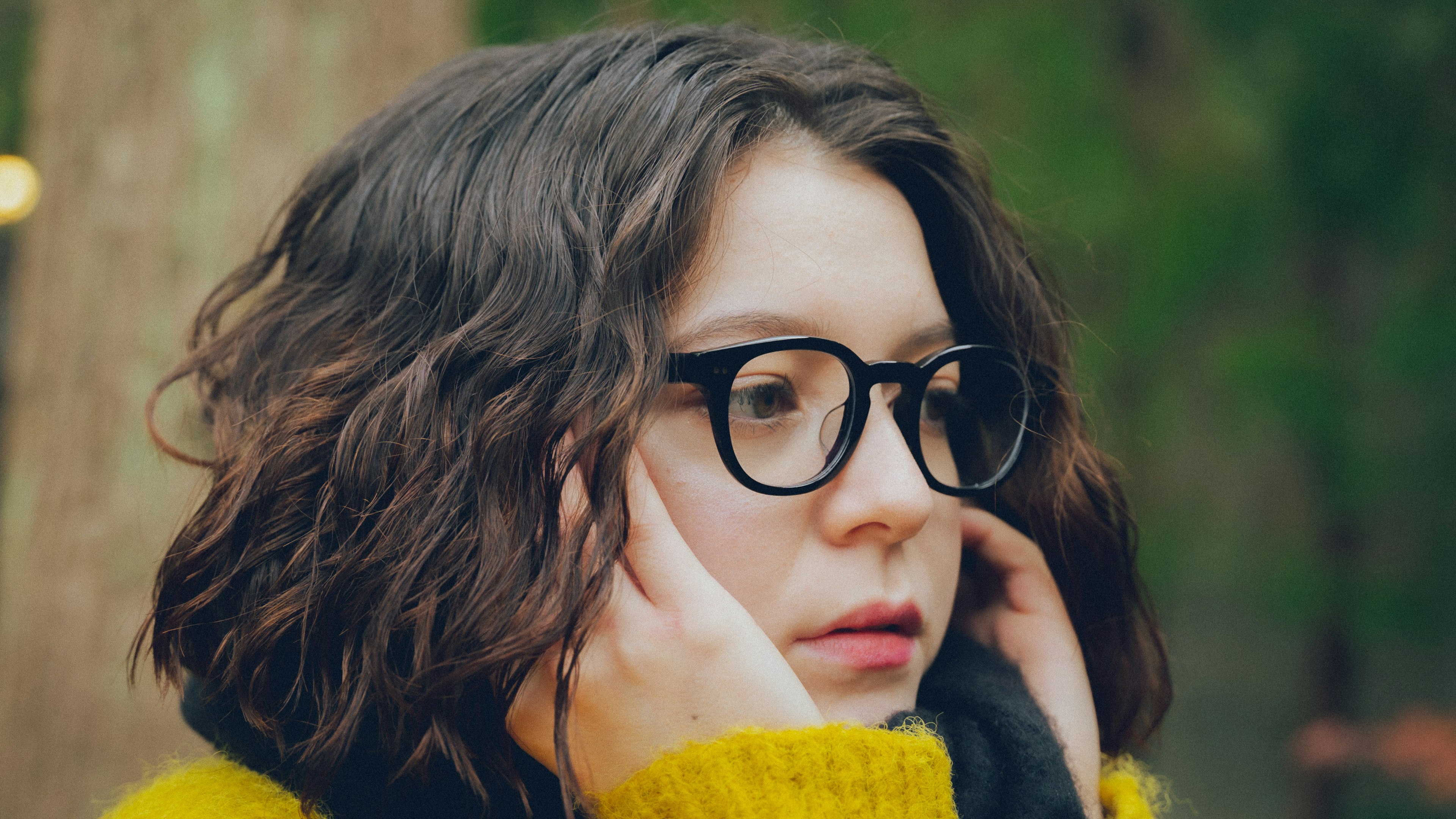 Perempuan muda dengan rambut keriting mengenakan kacamata menyentuh wajahnya di luar ruangan