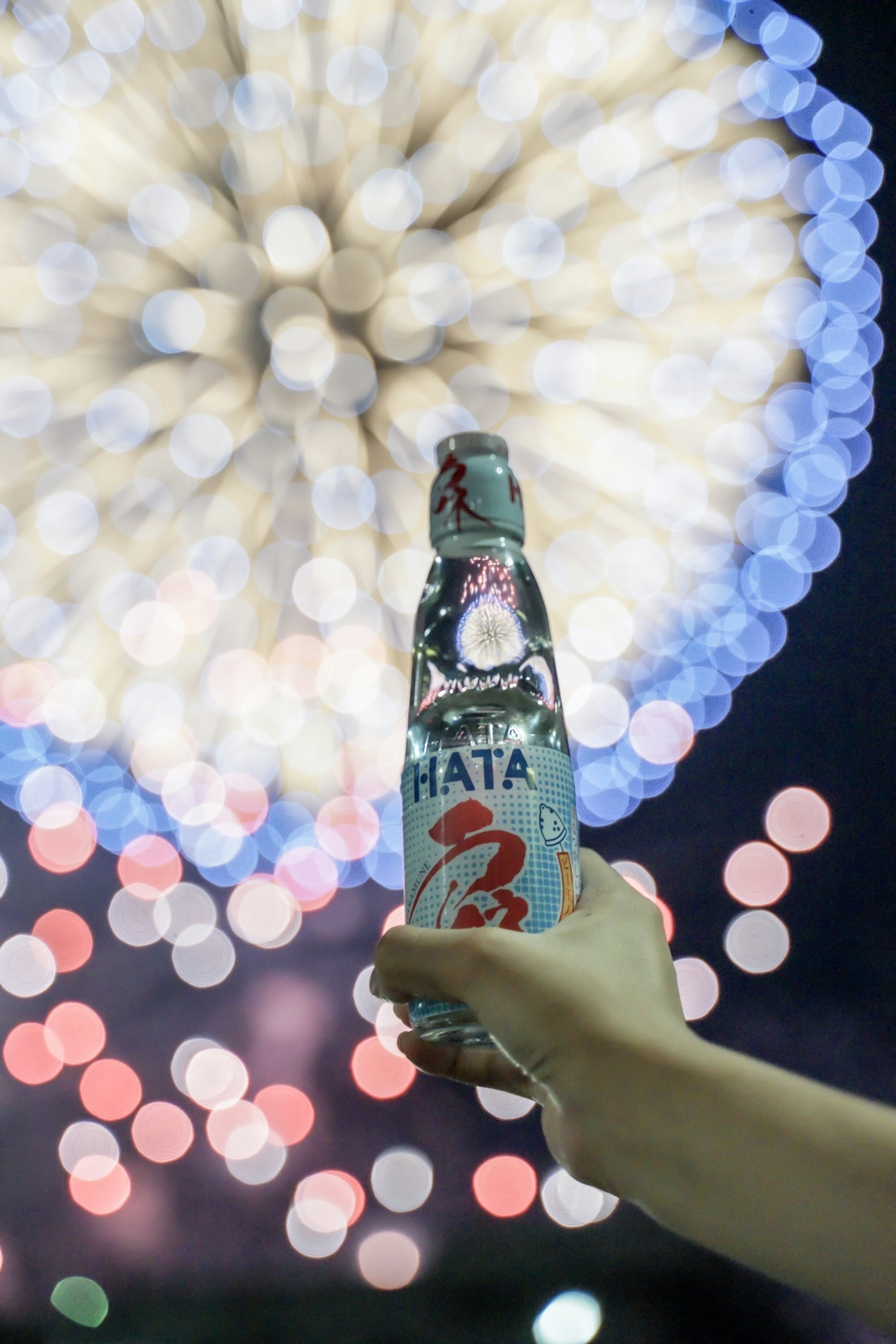 Mano sosteniendo una botella HATA con fuegos artificiales de fondo