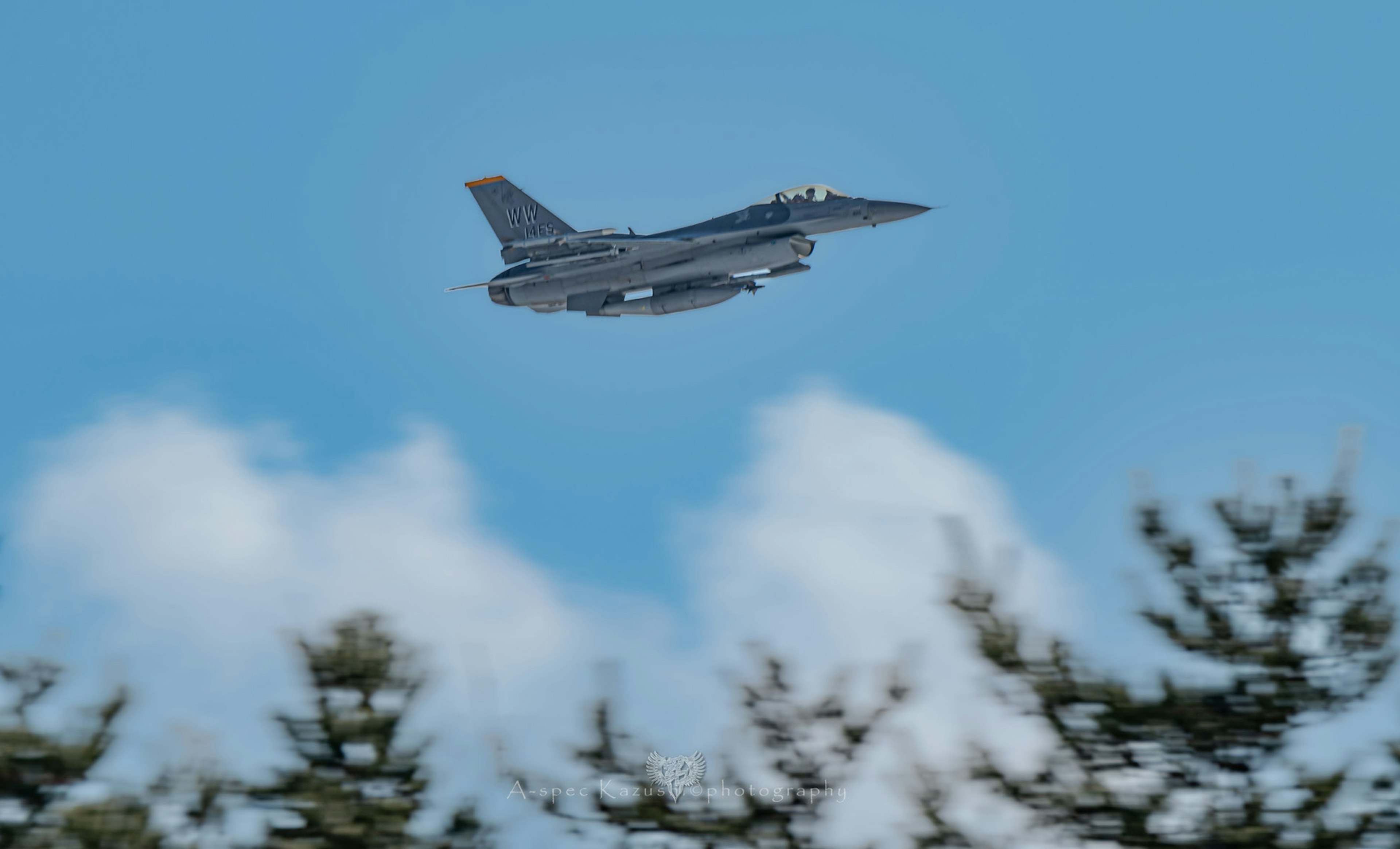 空を飛ぶ戦闘機の写真、青い空と白い雲、緑の木々が背景にある