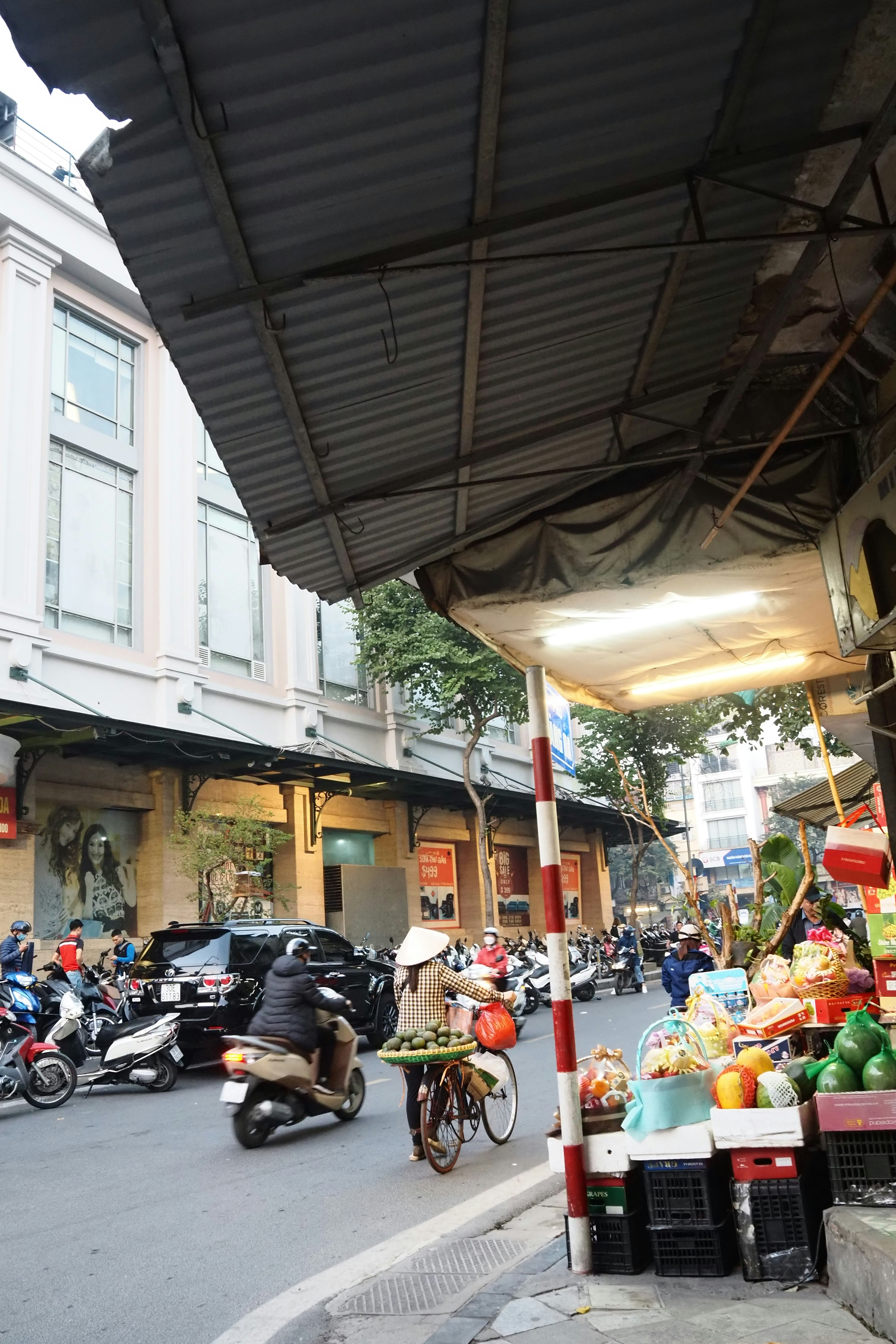 Bustling street market corner featuring fruits and vegetables with motorbikes passing