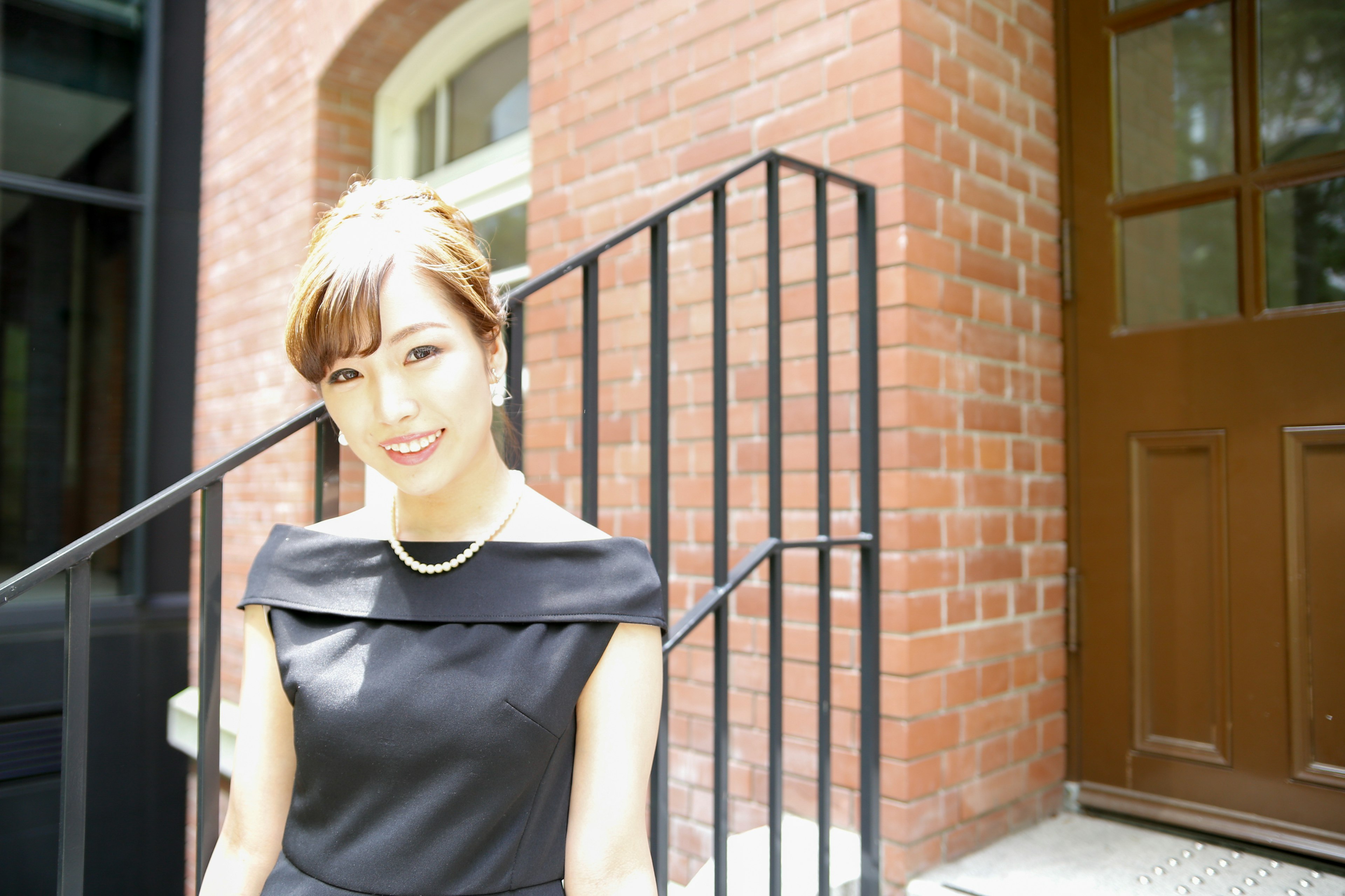 A woman in a black dress standing in front of stairs smiling casually Background features a red brick wall and door