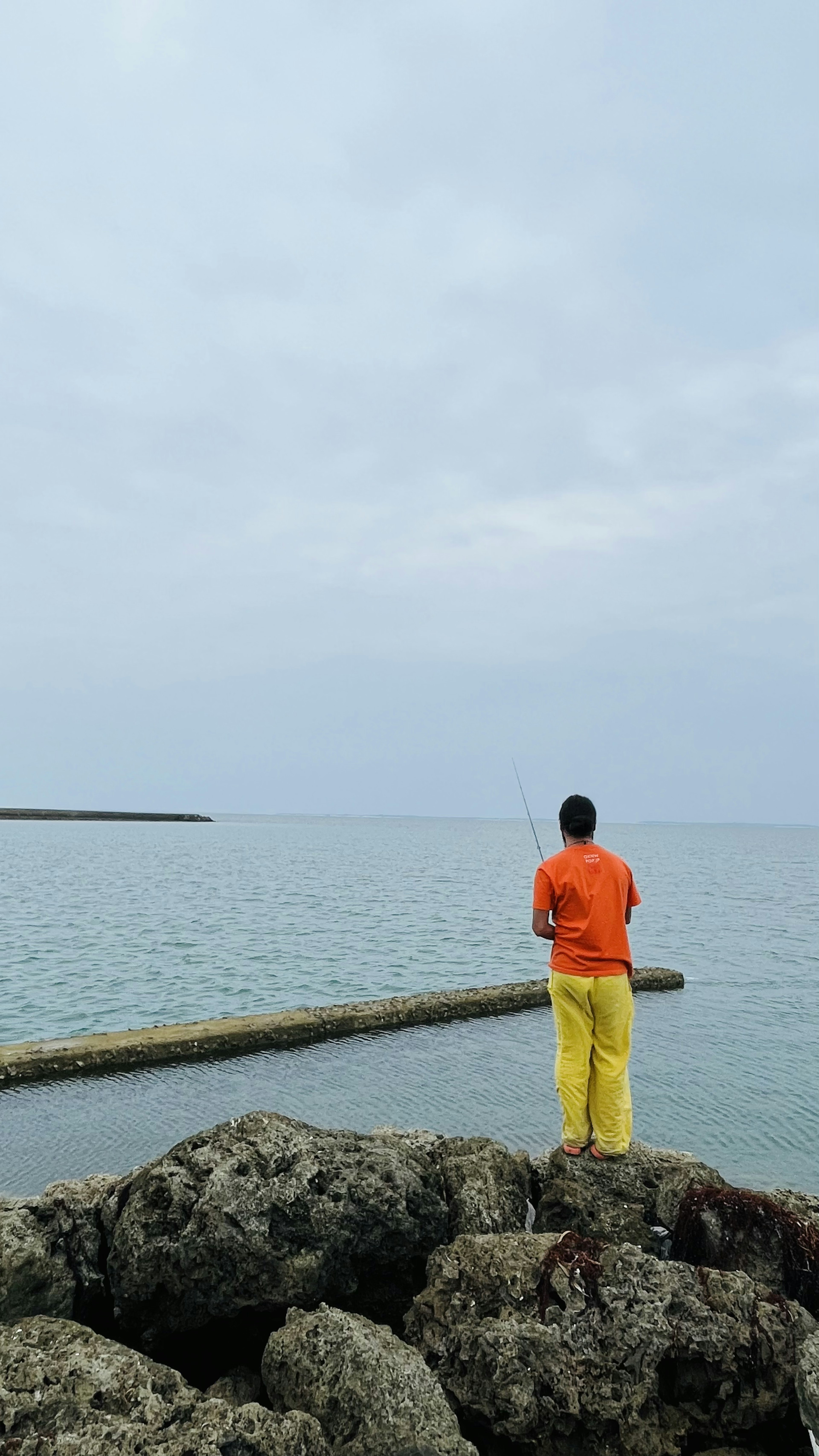 A man in an orange shirt and yellow pants standing on rocks looking at the sea