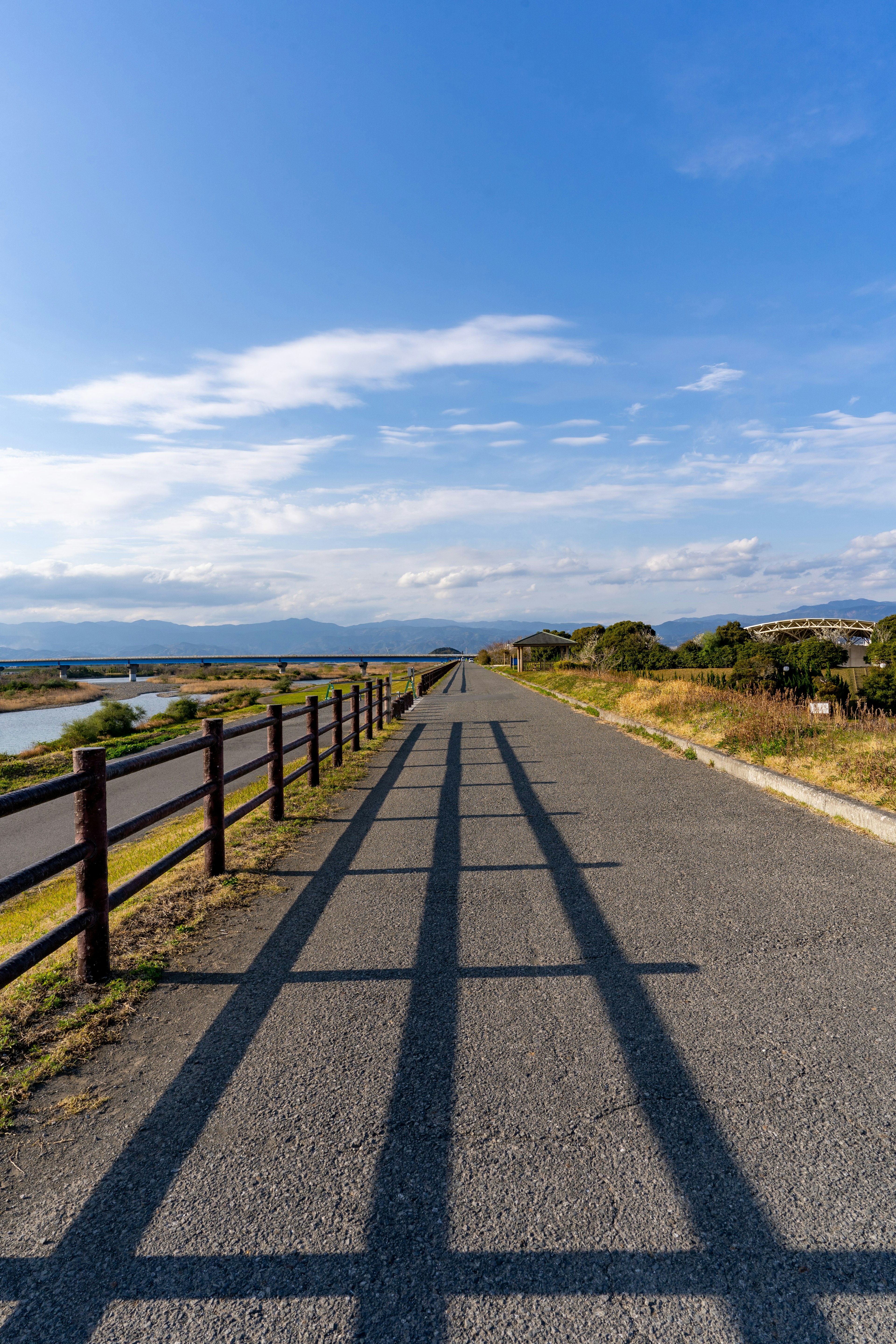 Paved path under blue sky with long shadows from the fence