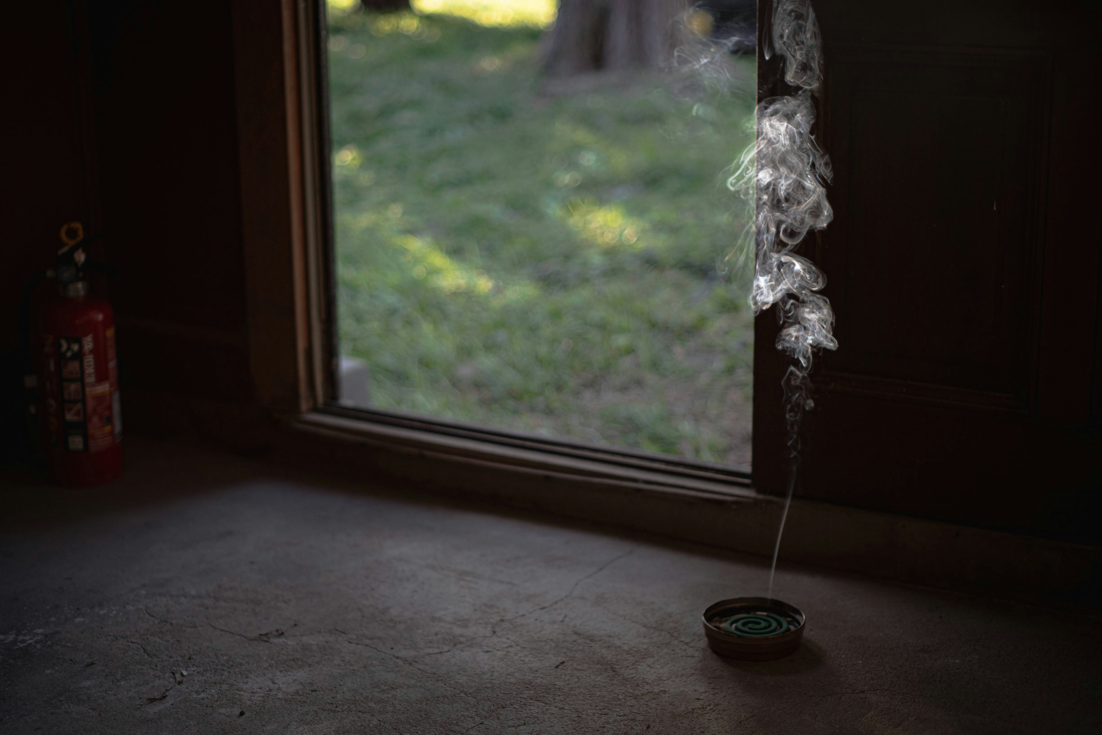 Interior view showing smoke drifting from an incense stick near a window with a green outdoor background