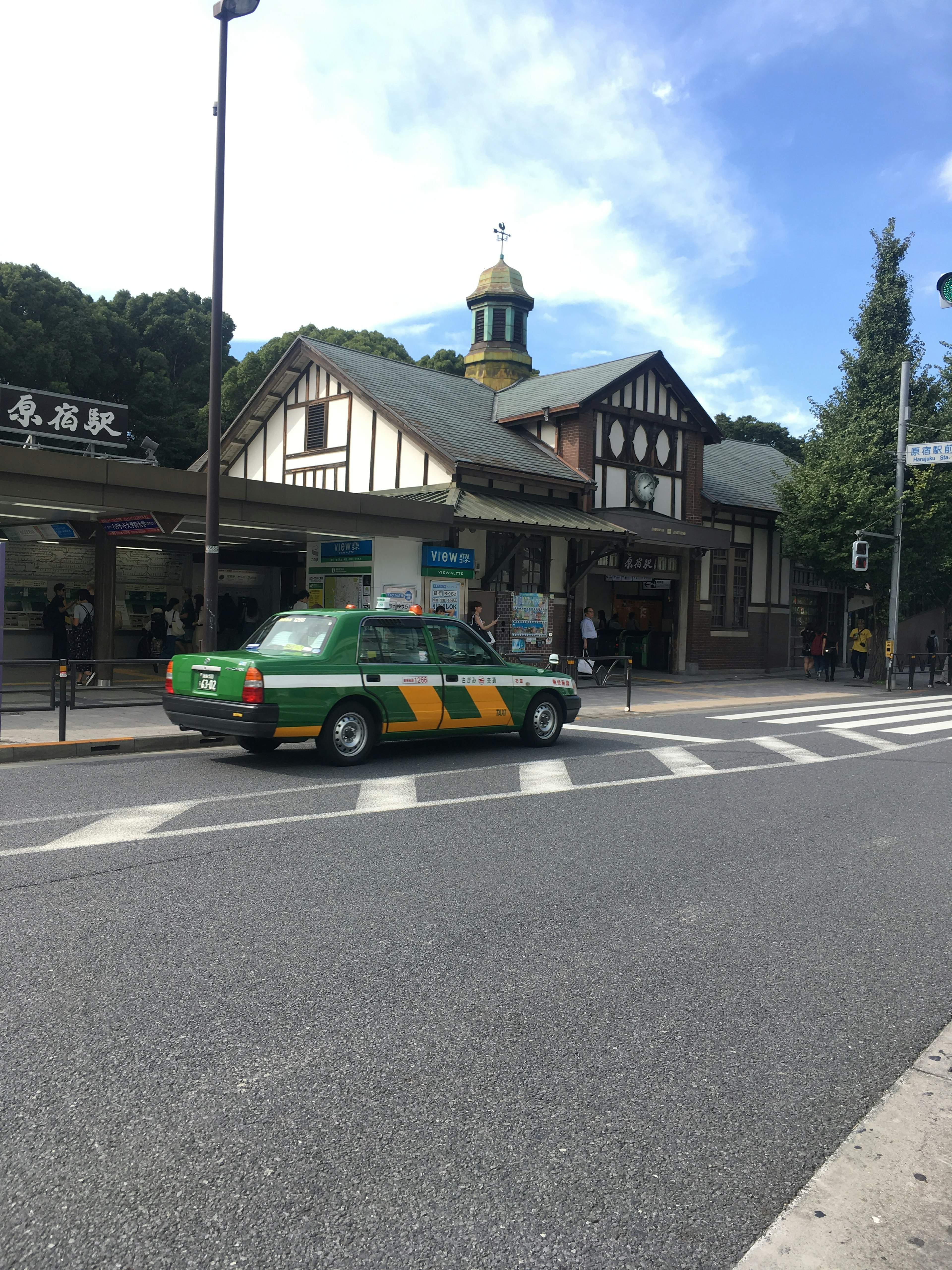 Taxi màu xanh lá cây và vàng đang chạy trên đường với một tòa nhà ga ở phía sau