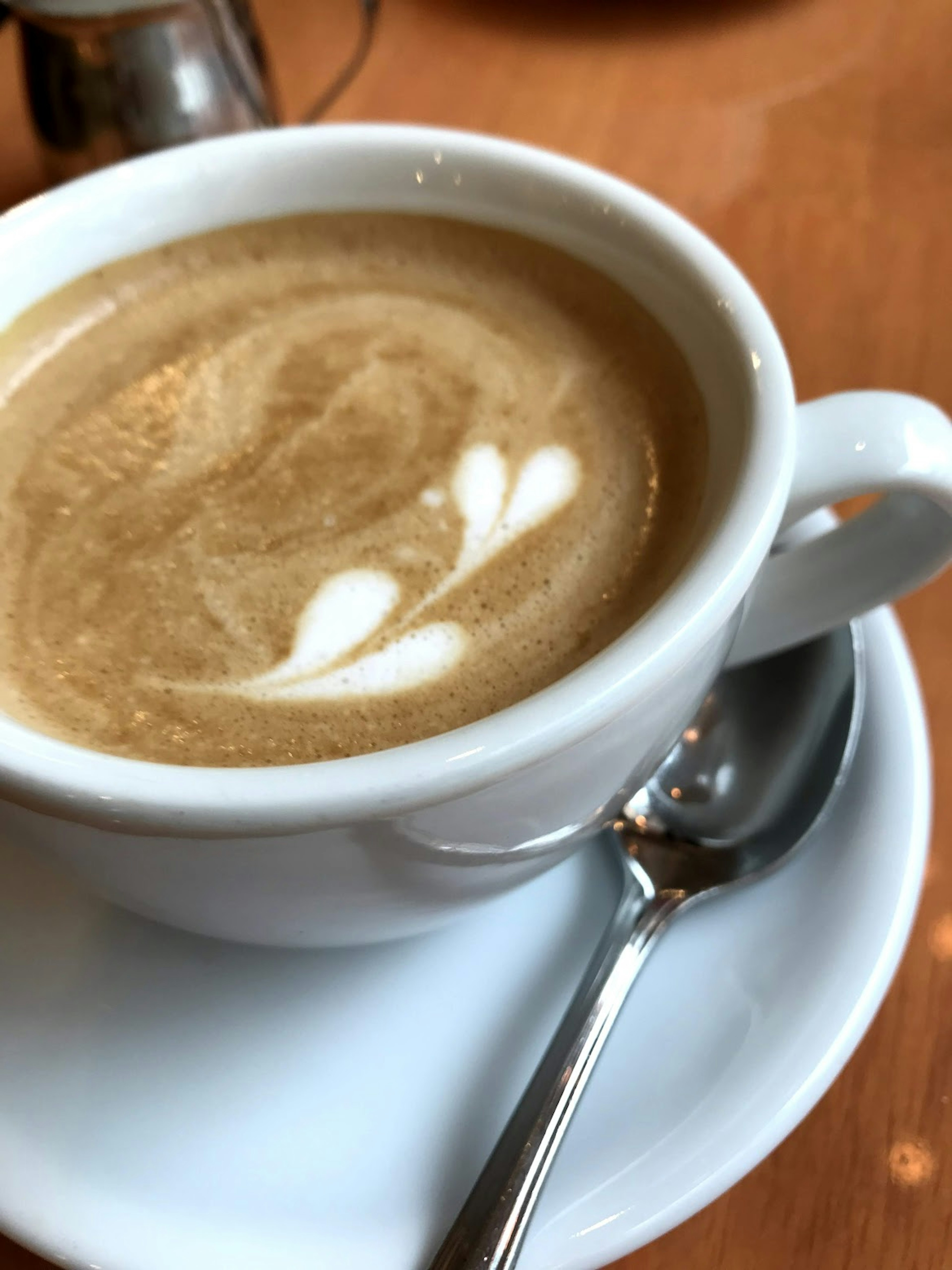 A cup of latte with latte art on a saucer and a spoon beside it