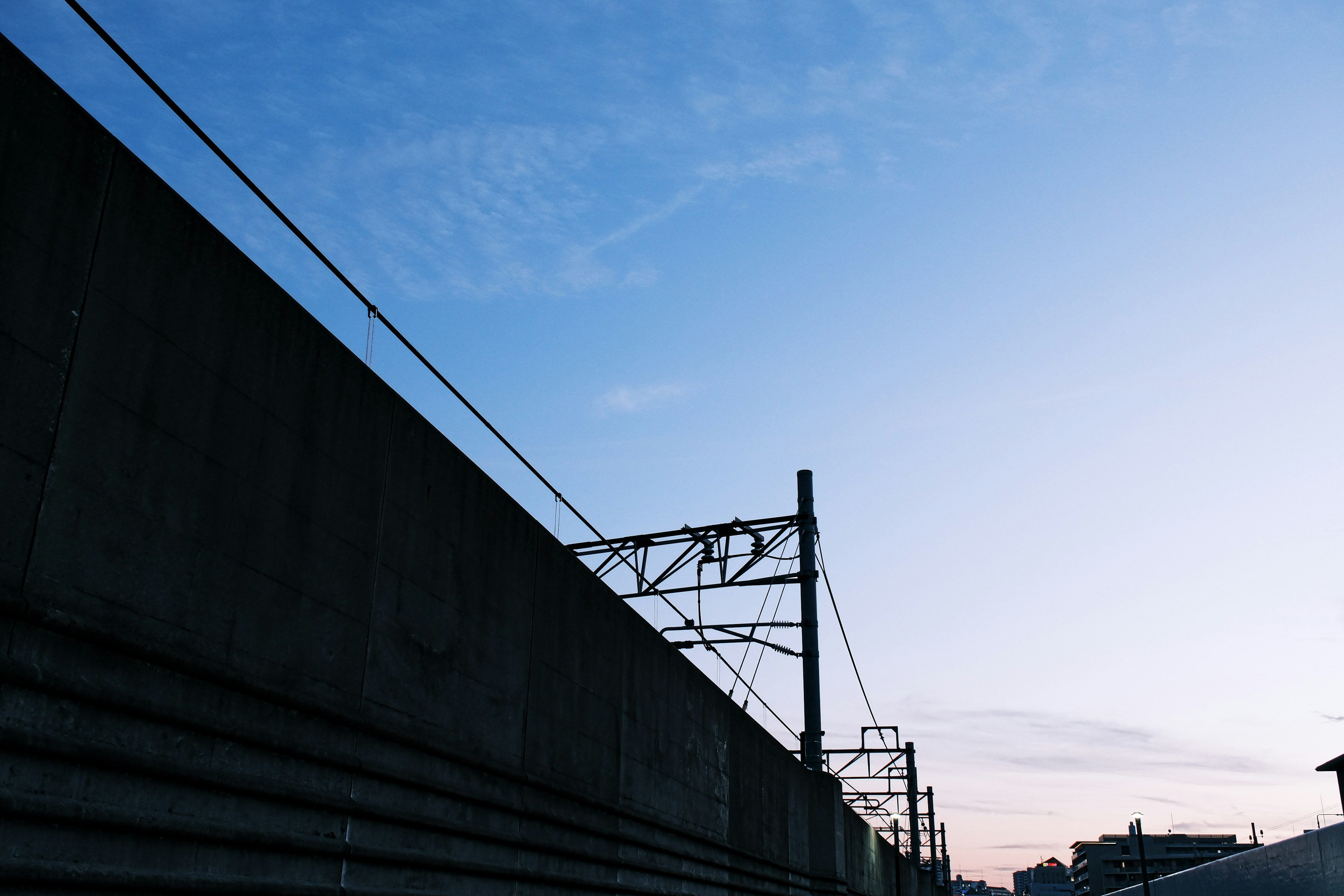 Muro de concreto con cables aéreos de tren bajo un cielo azul al atardecer