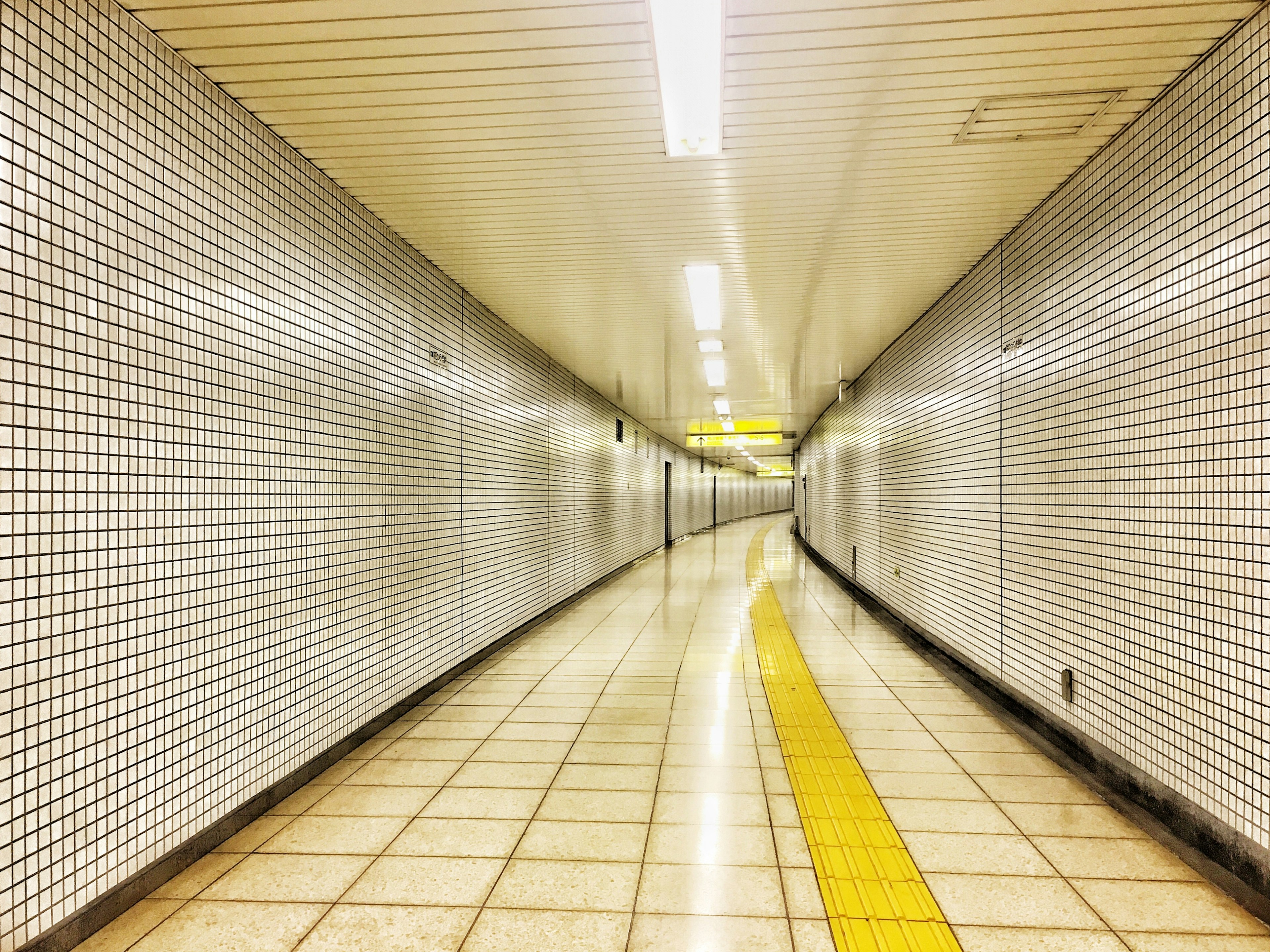 Pasillo subterráneo curvado con paredes y suelo de azulejos blancos que presenta una línea guía amarilla prominente