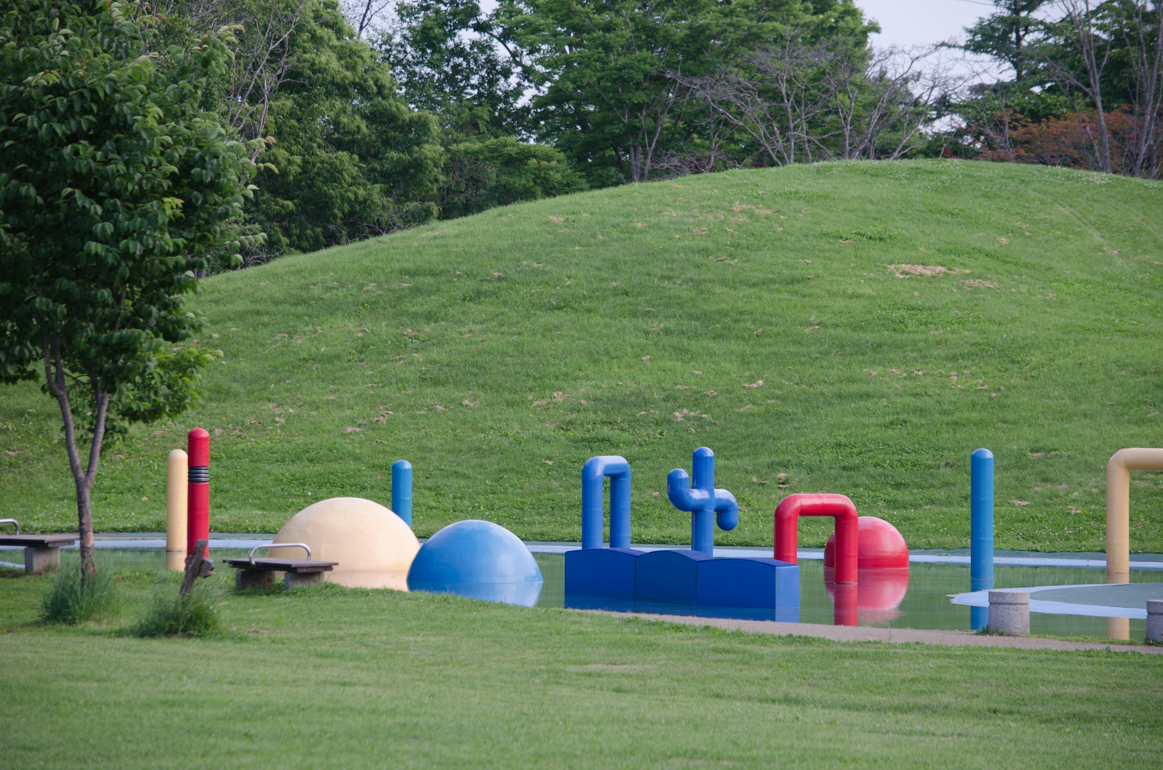 Colorful playground equipment in a park with a green hill and trees in the background