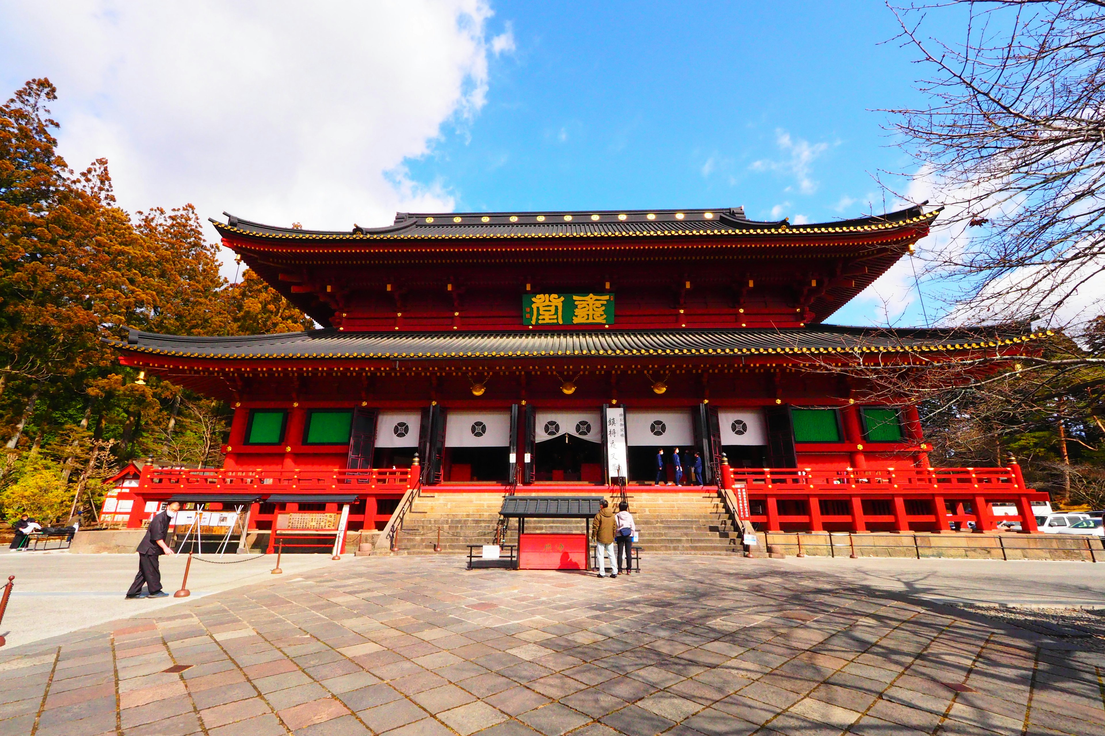 Vista frontal de un templo japonés tradicional con techo rojo cielo azul y nubes blancas rodeado de árboles verdes