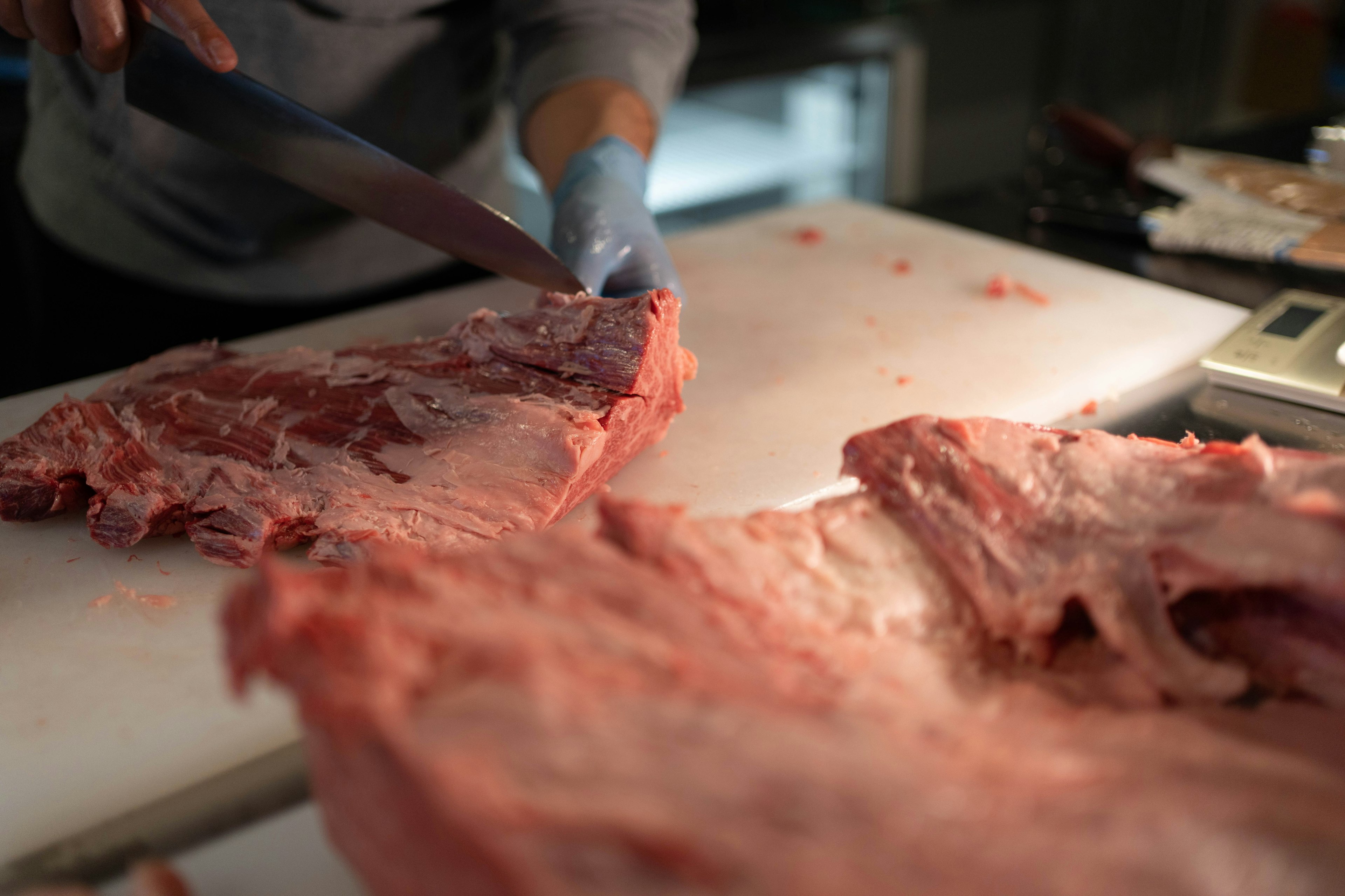 Gambar close-up tangan memotong daging dengan pisau sambil mengenakan sarung tangan biru