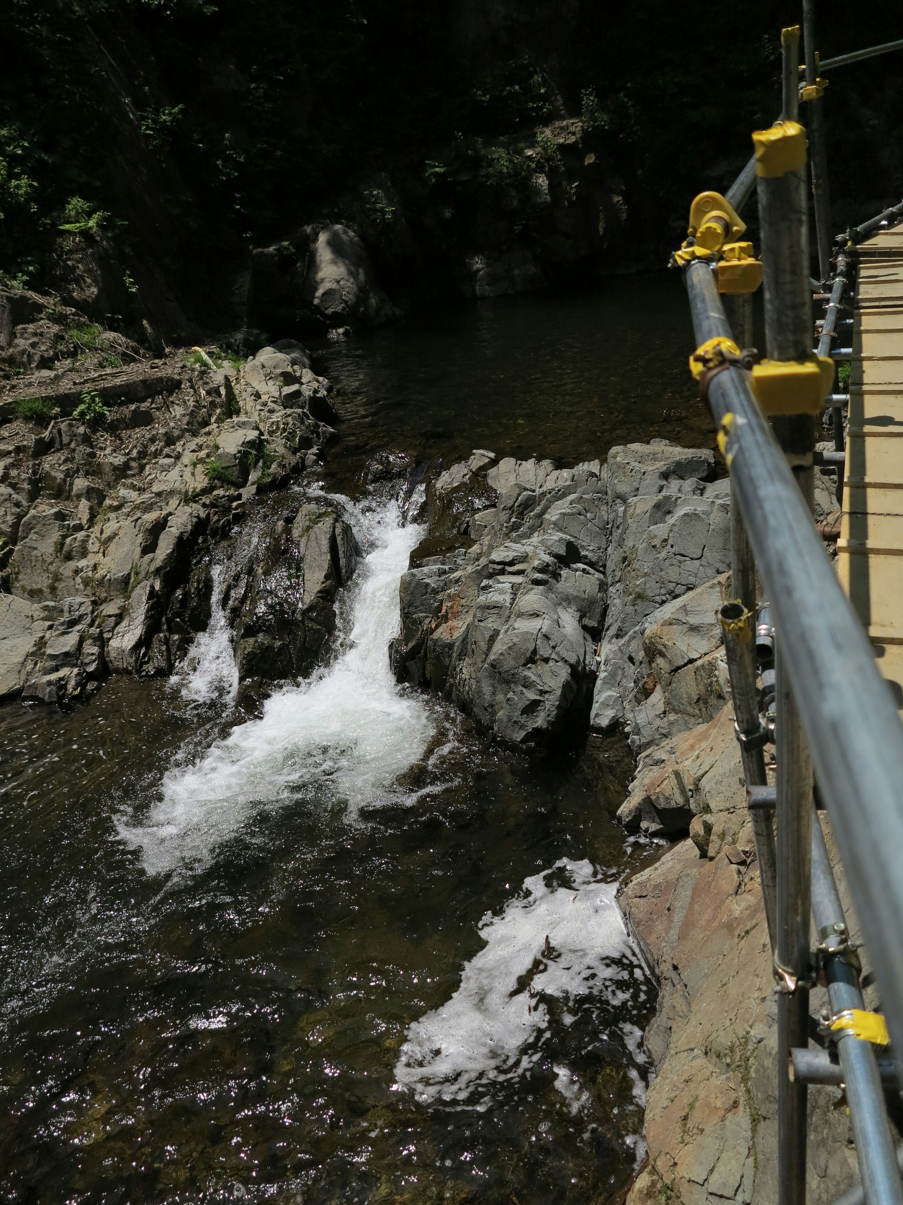 Vista panoramica di un torrente con rocce e un ponte con parapetto giallo