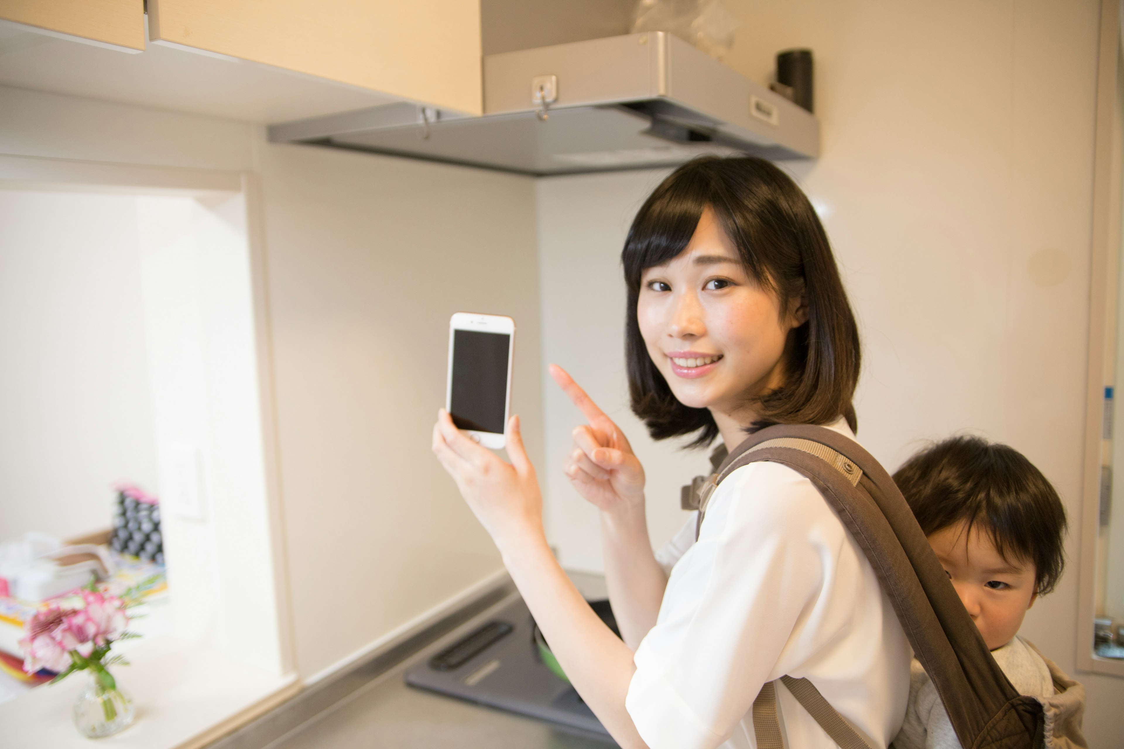 Woman in a kitchen holding a smartphone with a child behind her