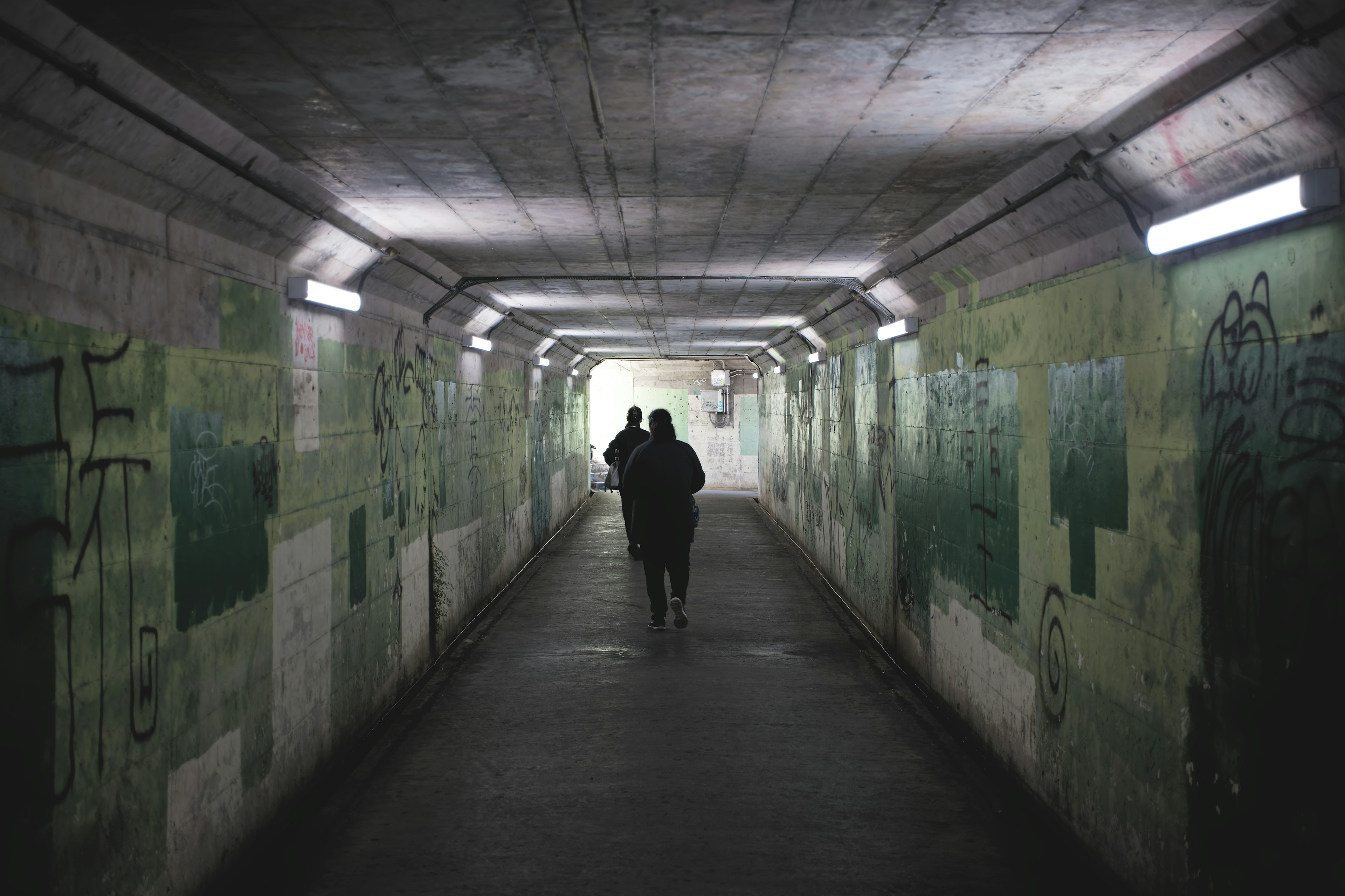 Zwei Figuren gehen in einem dunklen Tunnel mit grünen Wänden und Graffiti, beleuchtet von schwachem Licht