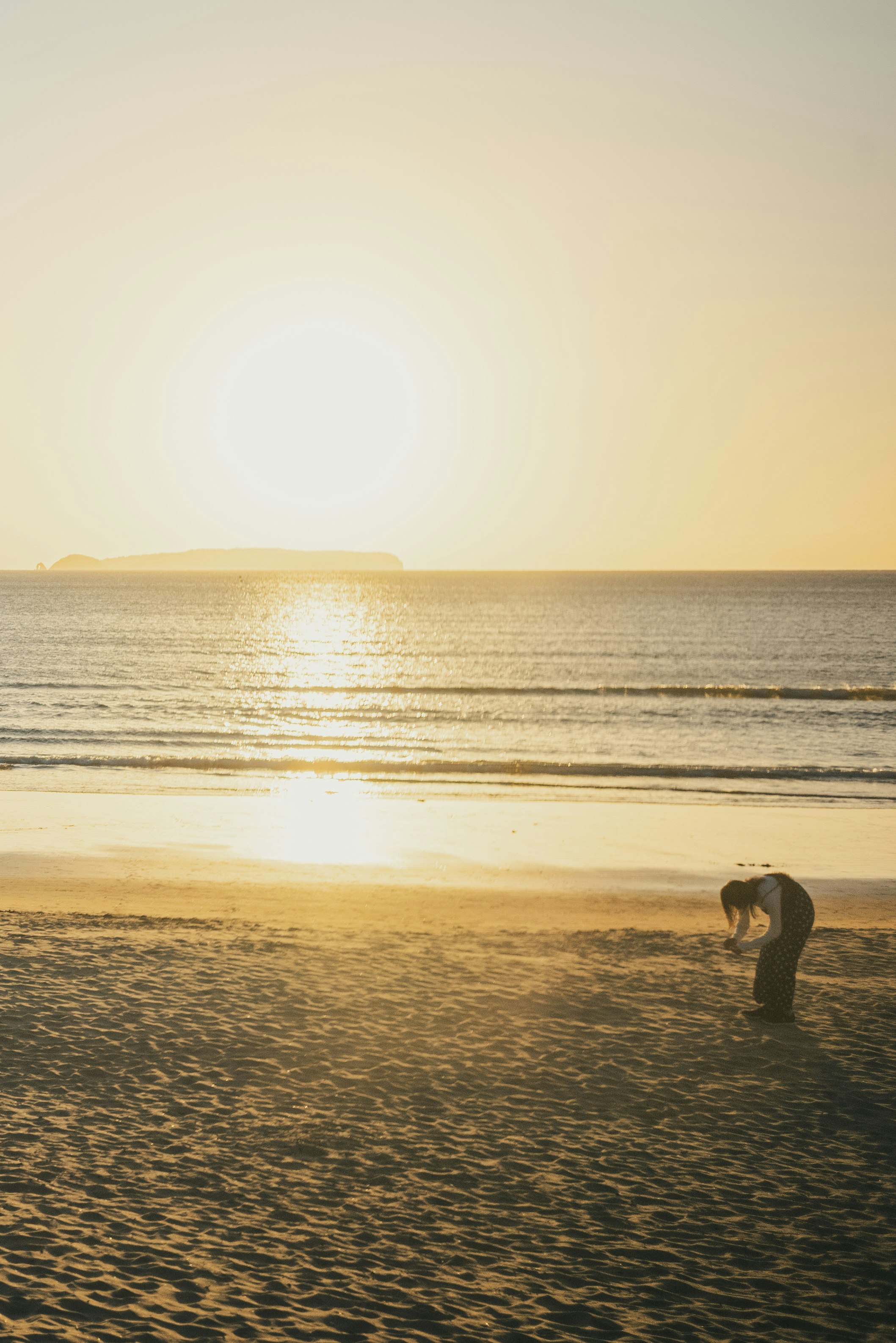 A person crouching on the sandy beach with the sunset in the background