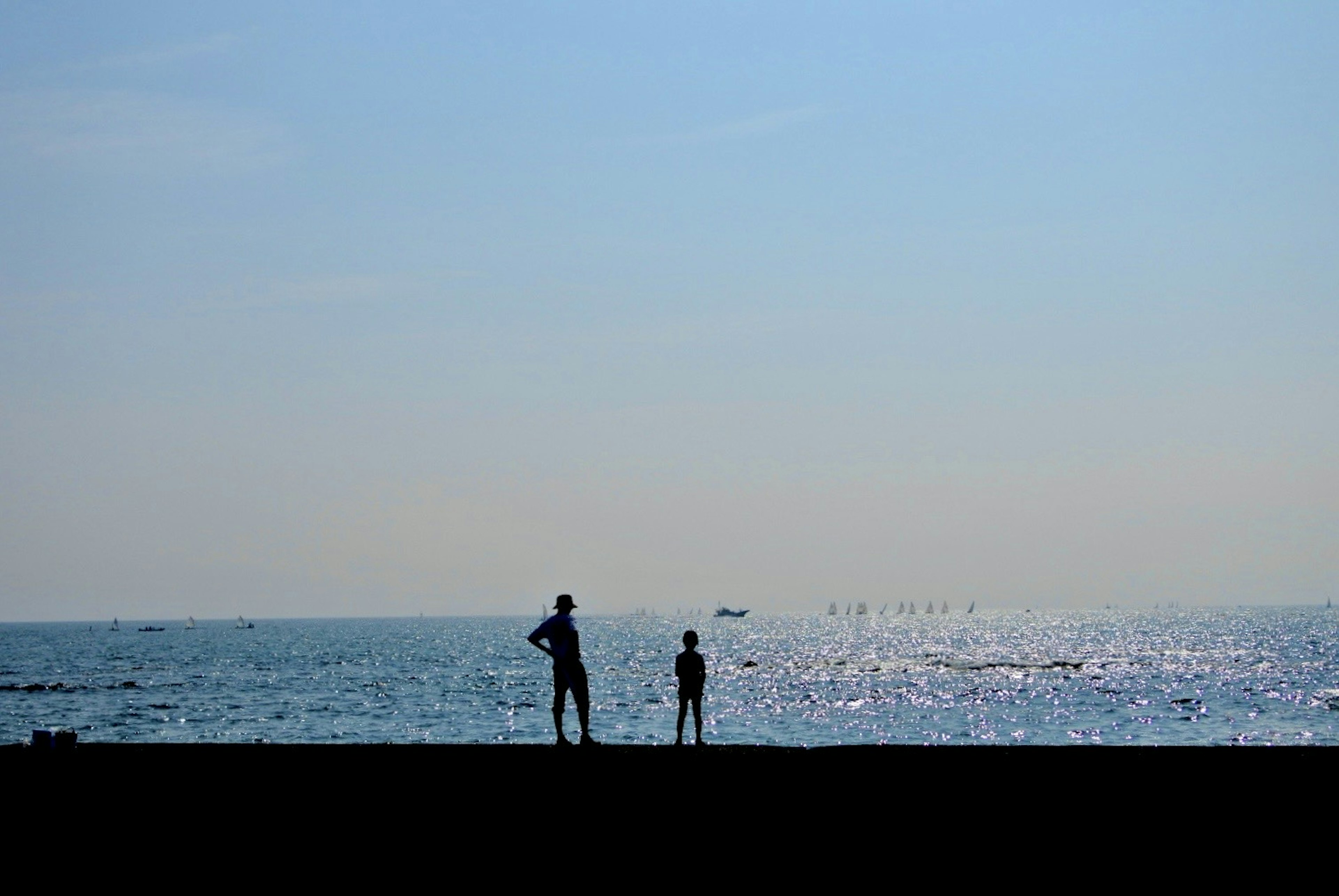 Silhouette eines Erwachsenen und eines Kindes am Meer mit blauem Himmel und schimmerndem Ozean