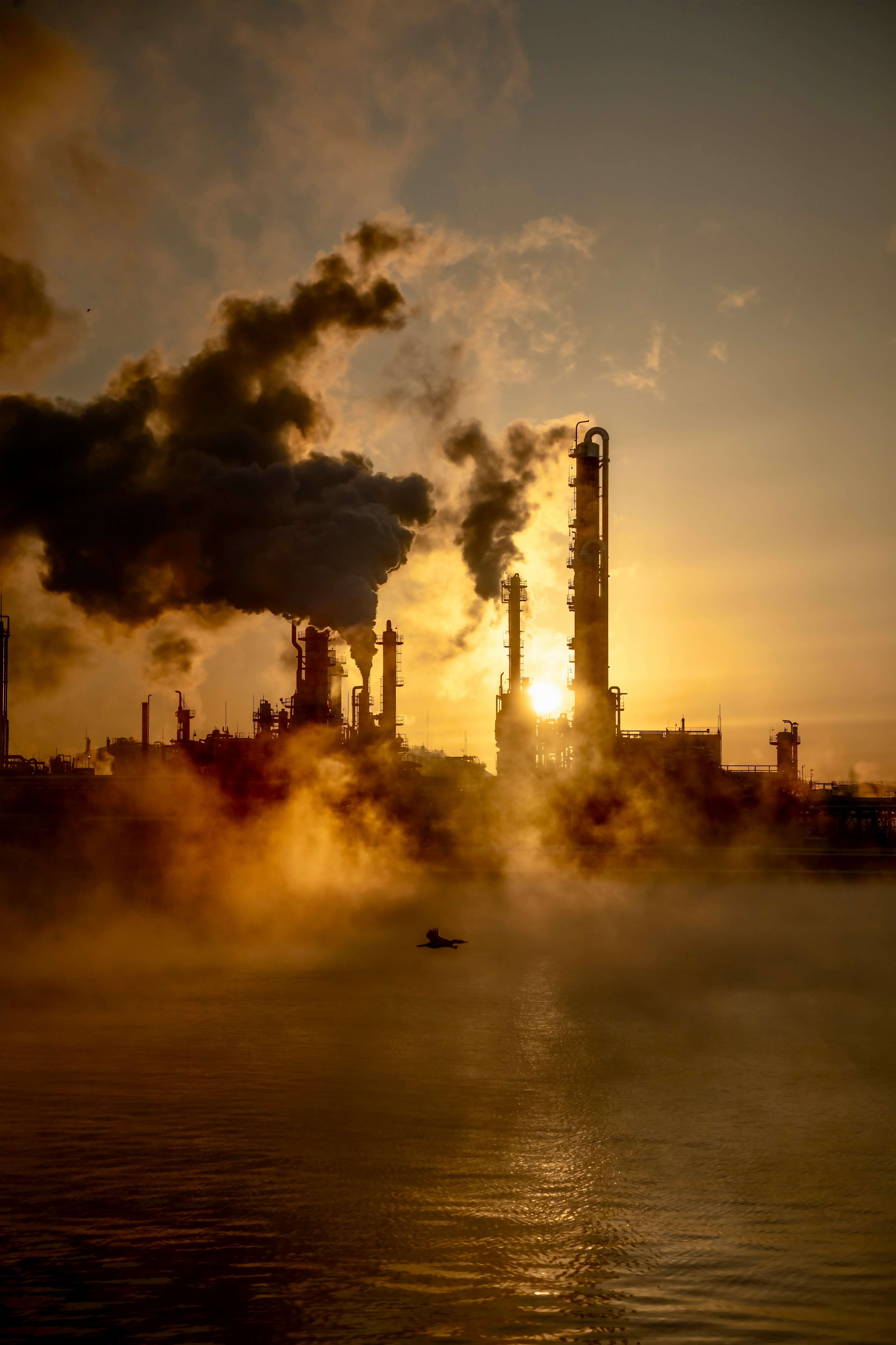 Silhouette d'usines émettant de la fumée au coucher du soleil avec des reflets sur l'eau