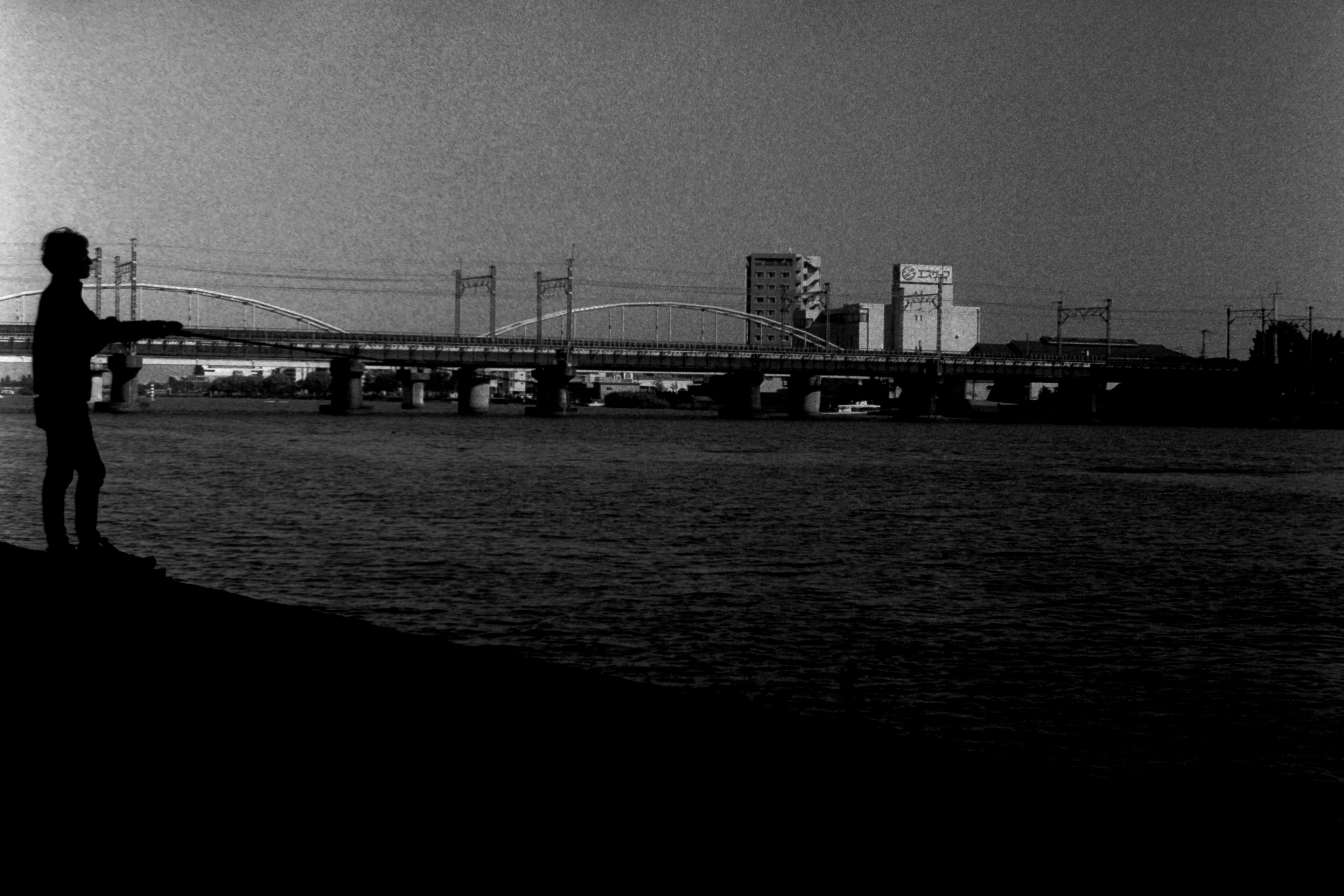 Silhouette einer Person am Ufer des Flusses mit entfernten Gebäuden in einer monochromen Landschaft