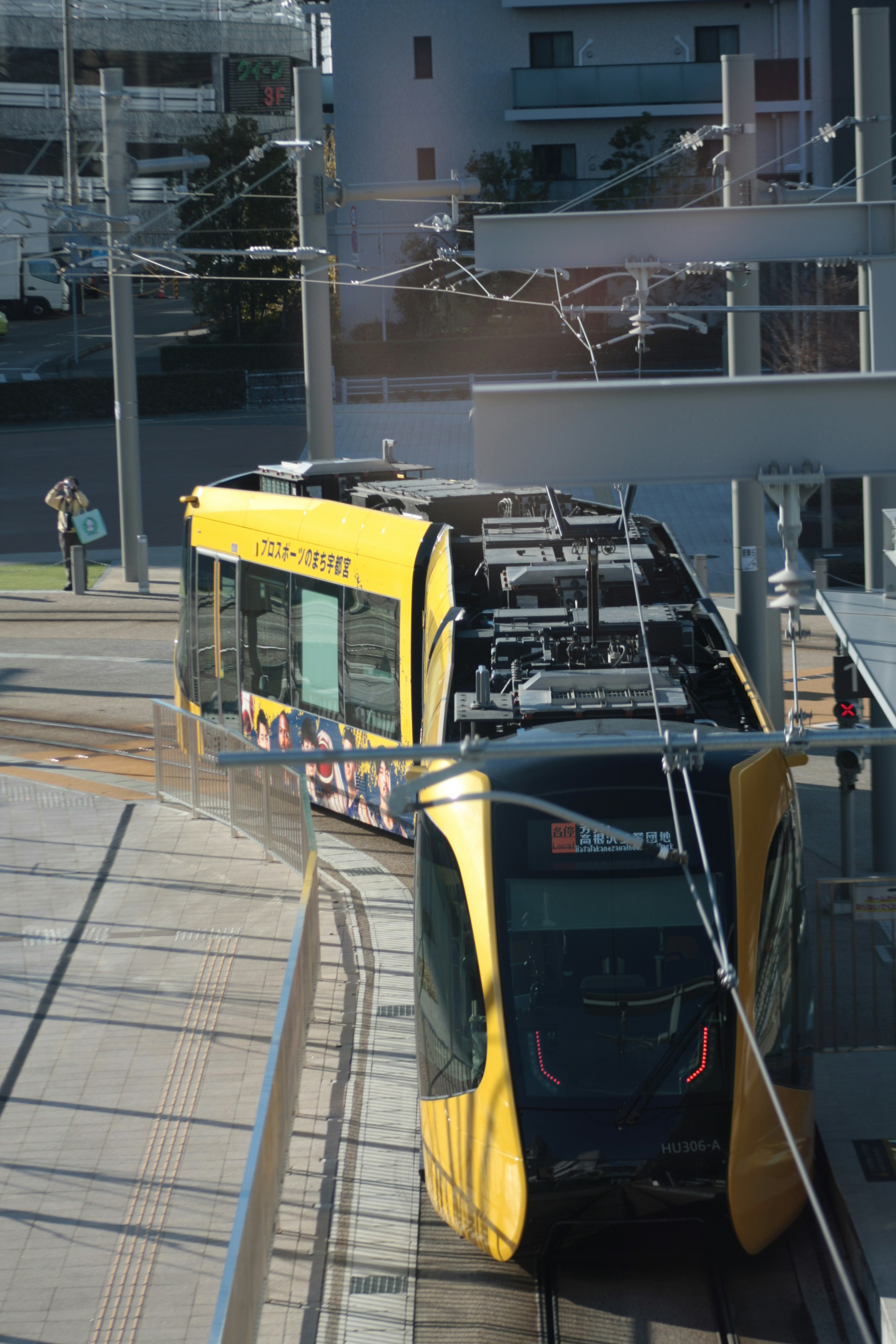 Tram kuning berhenti di stasiun dengan gedung modern di latar belakang