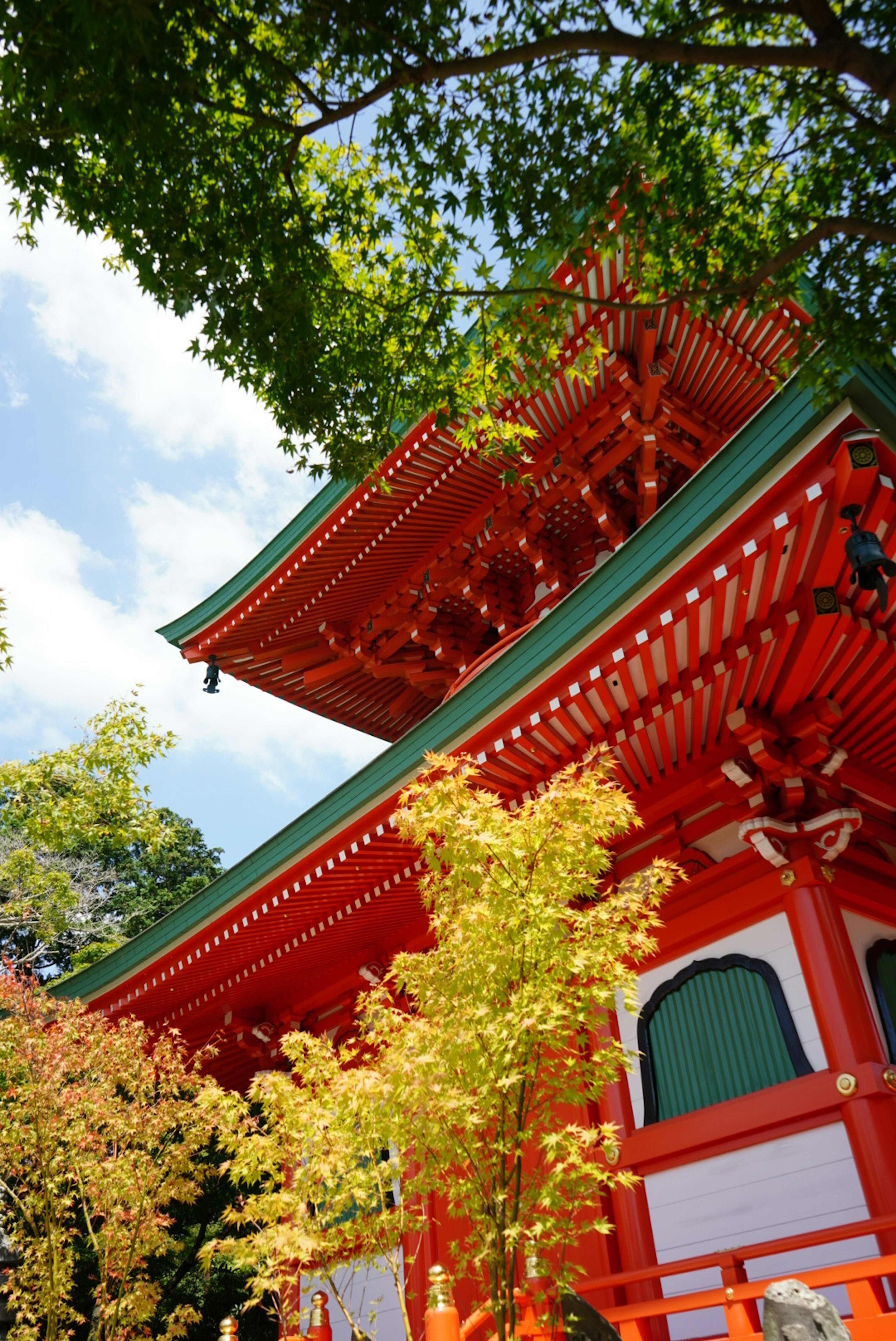 美麗的日本寺廟建築，紅色牆壁和綠色屋頂，四周環繞著綠色樹木和黃色葉子