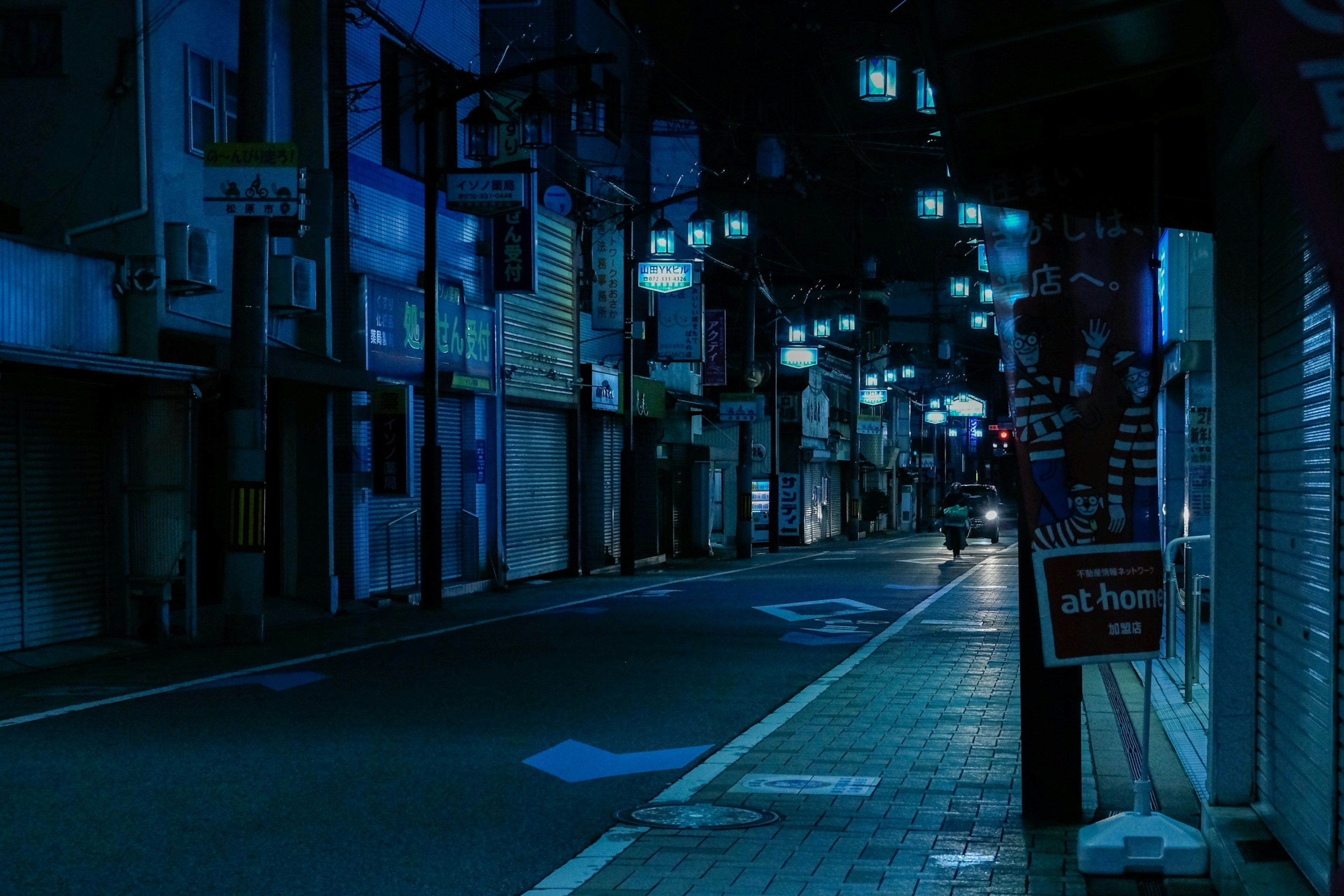 静かな夜の街並み　青い照明に照らされた通り　閉まった店舗と人影