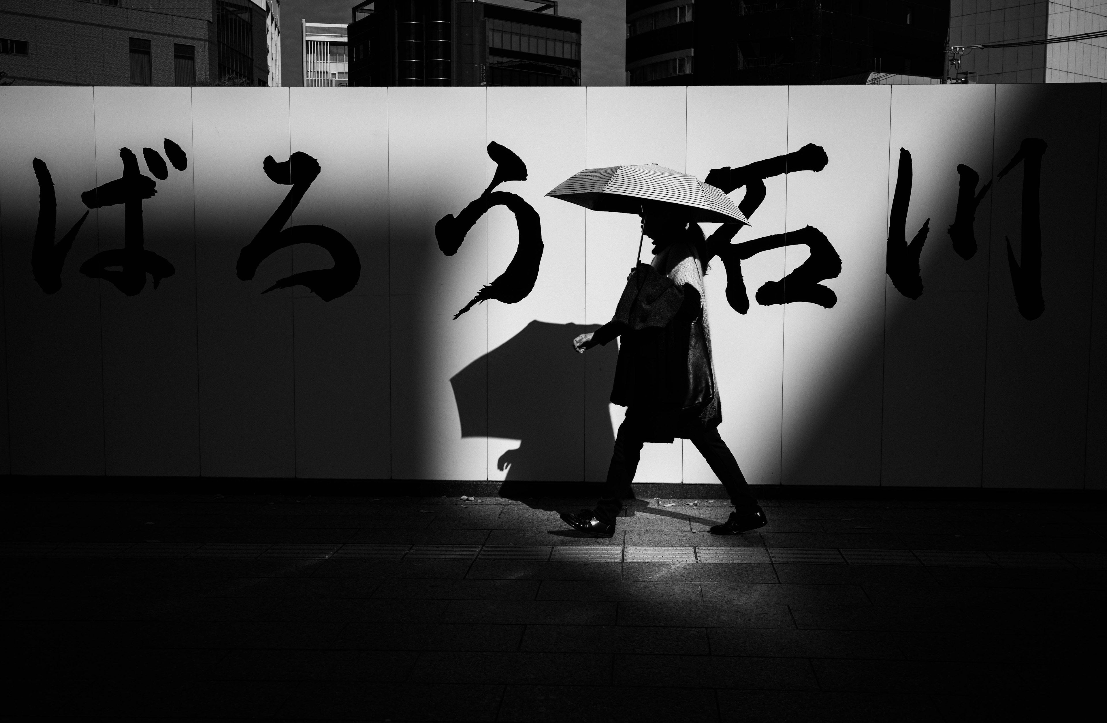 Silueta de una persona caminando con un paraguas negro frente a una pared con caligrafía japonesa