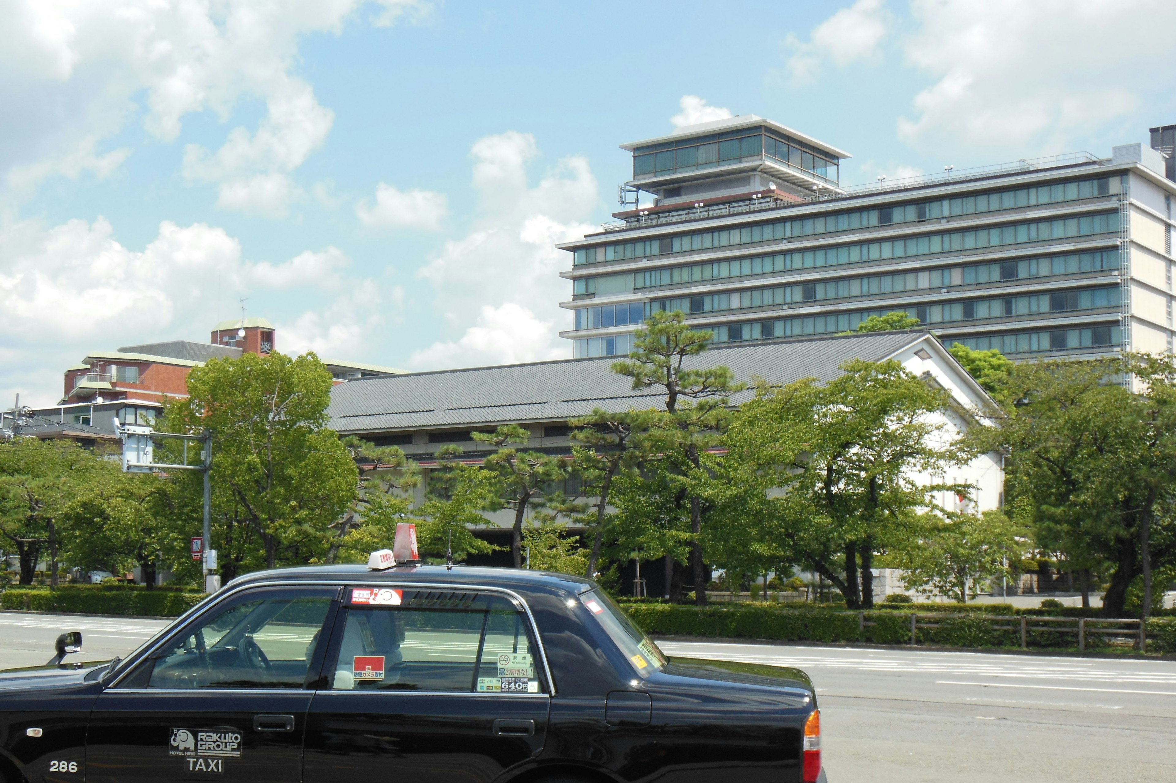 Sebuah taksi di depan gedung modern dengan pohon dan langit biru