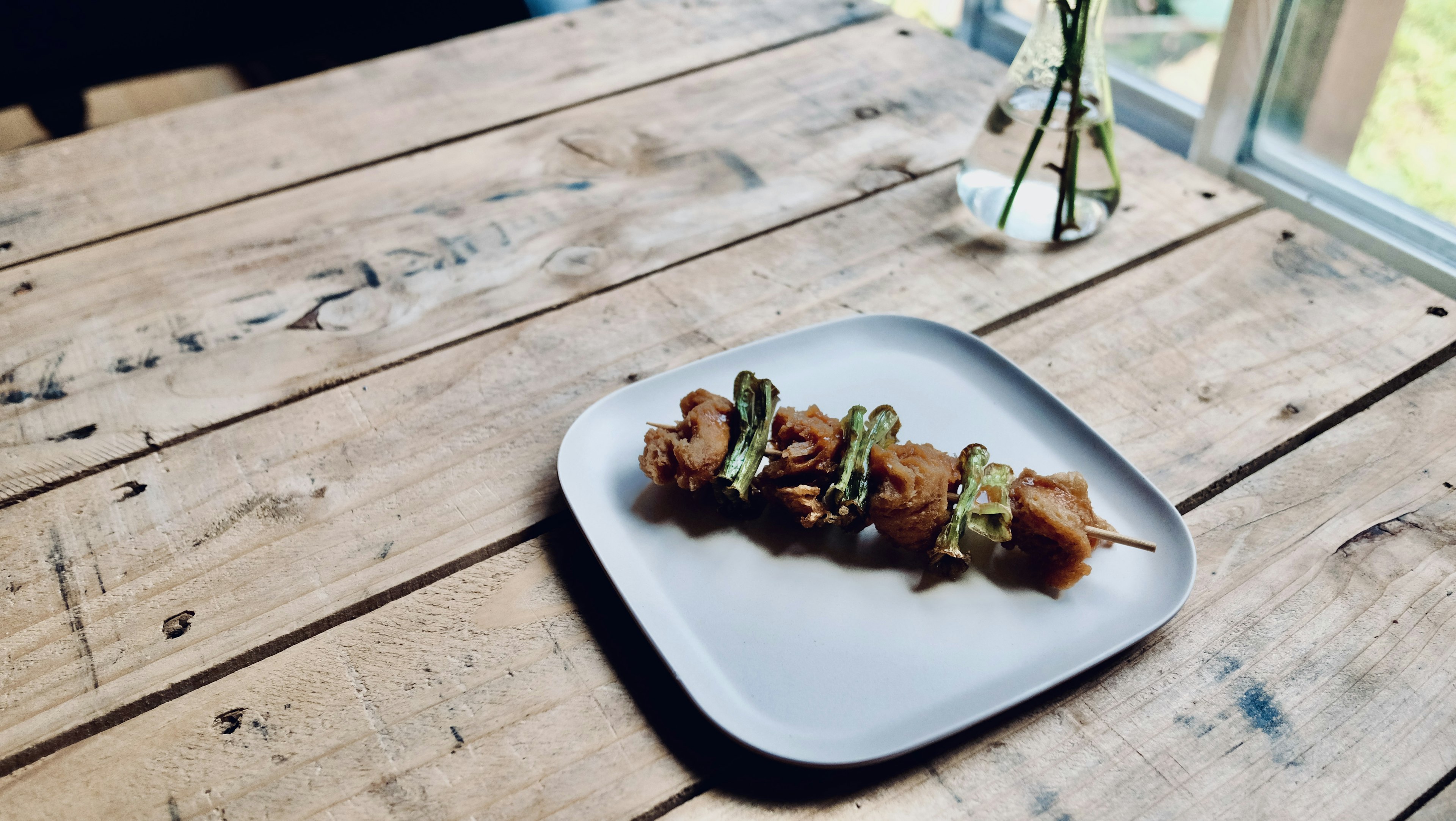 Skewers served on a plate on a wooden table with a small vase