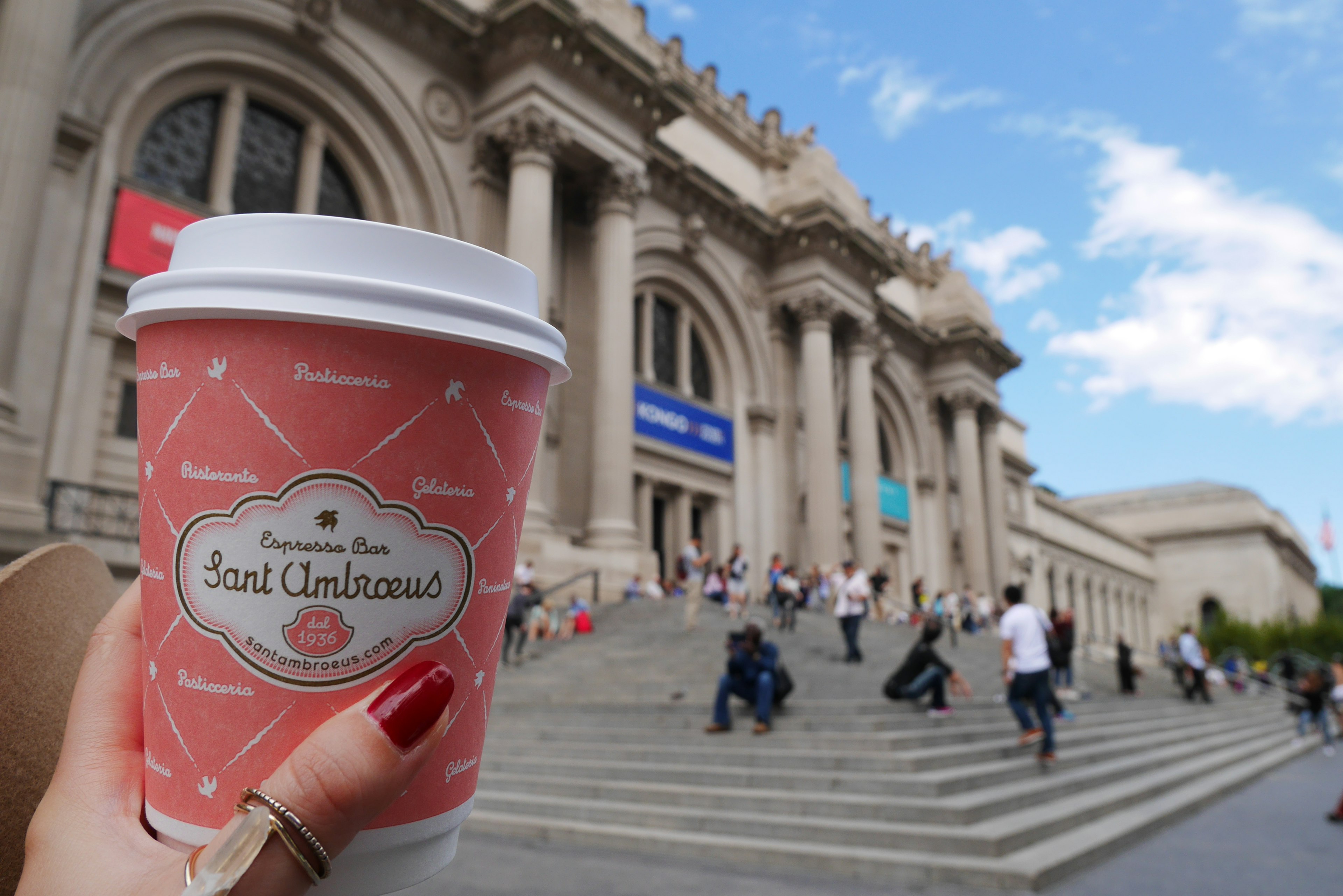 Mano sosteniendo una taza de café rosa frente al Museo Metropolitano de Arte