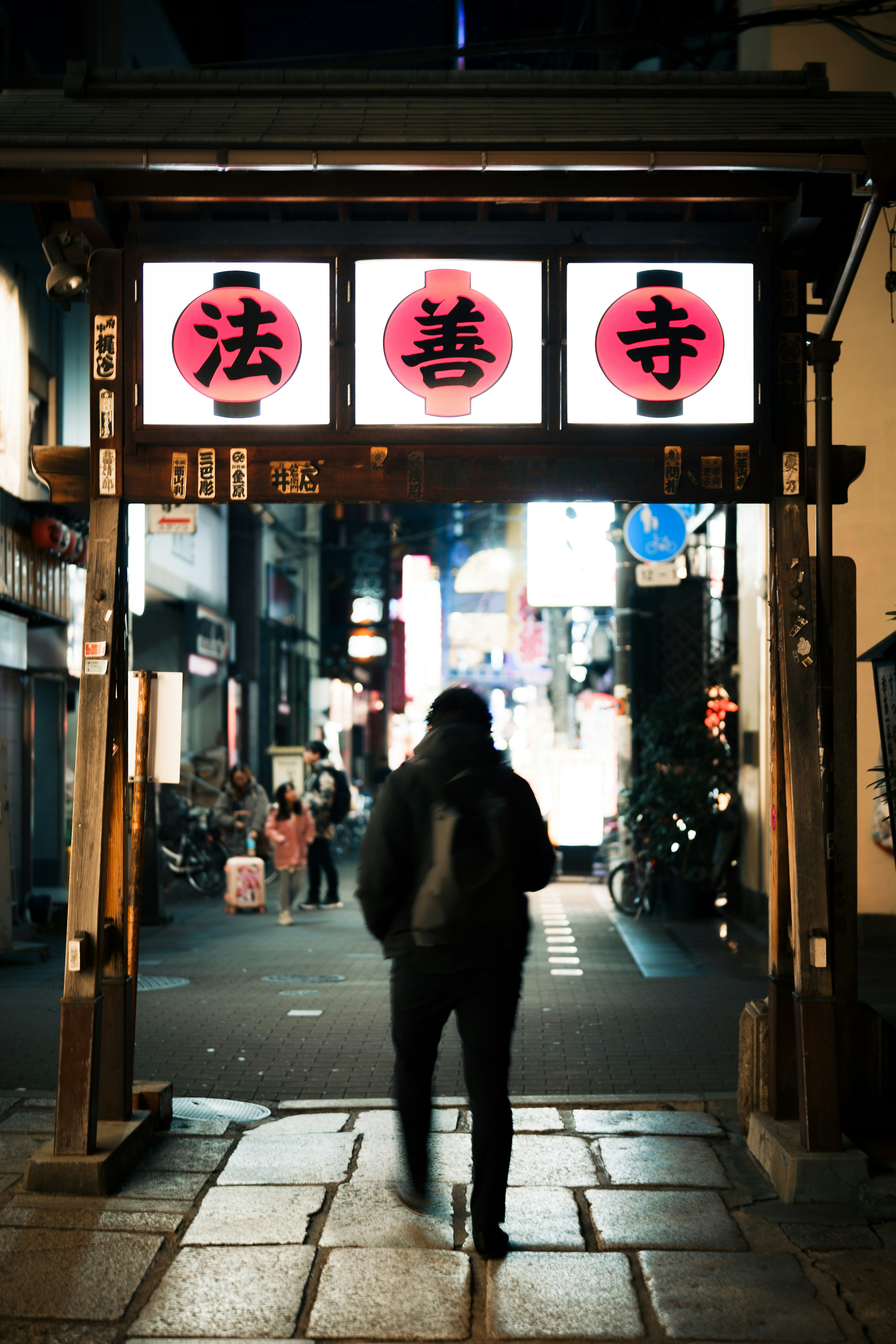 夜の街並みに立つ赤い提灯の門を通り抜ける人物