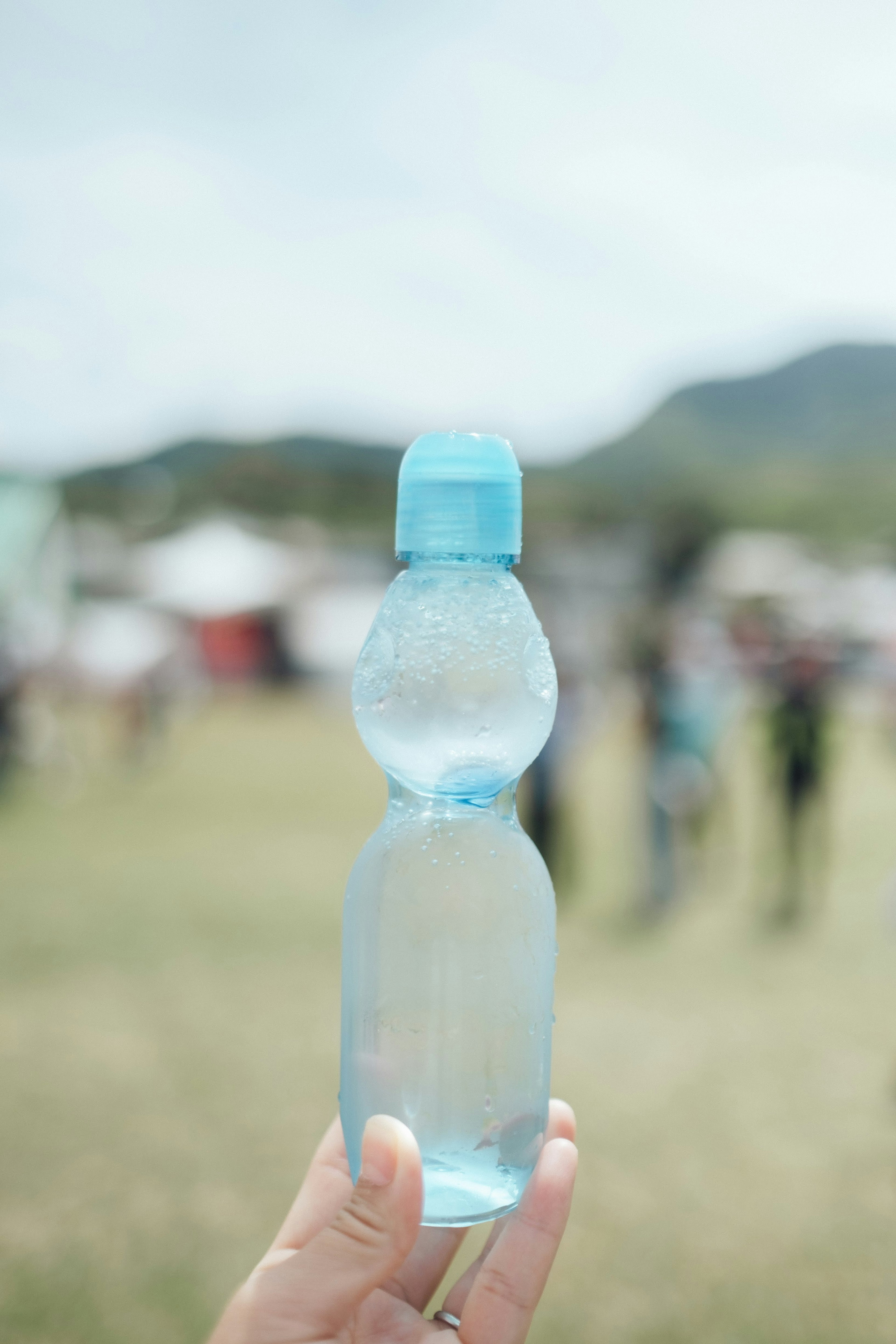 Tangan memegang botol air jernih dengan tutup biru dan orang-orang kabur di latar belakang