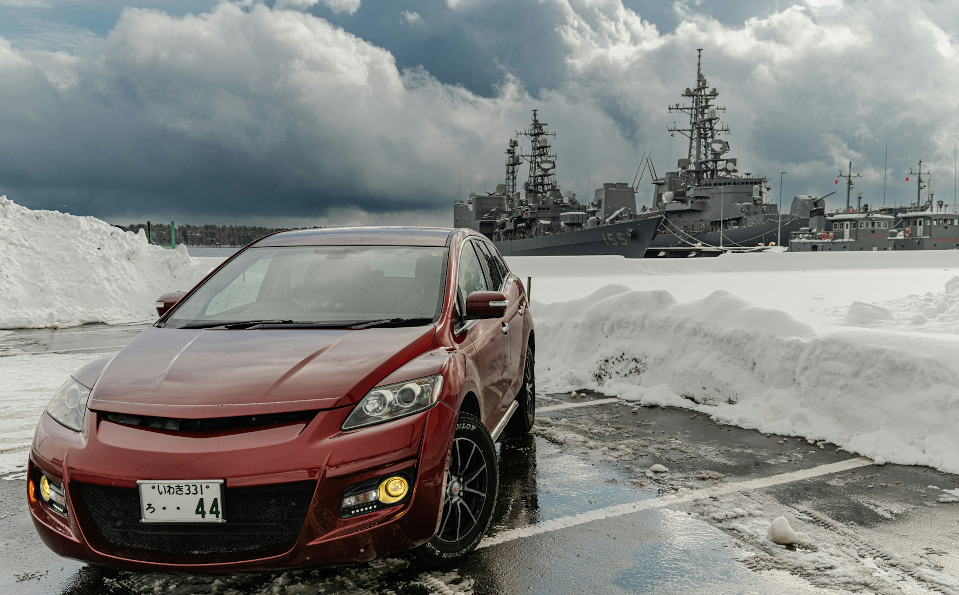 Red car parked in snowy lot with naval ships and dark clouds in the background