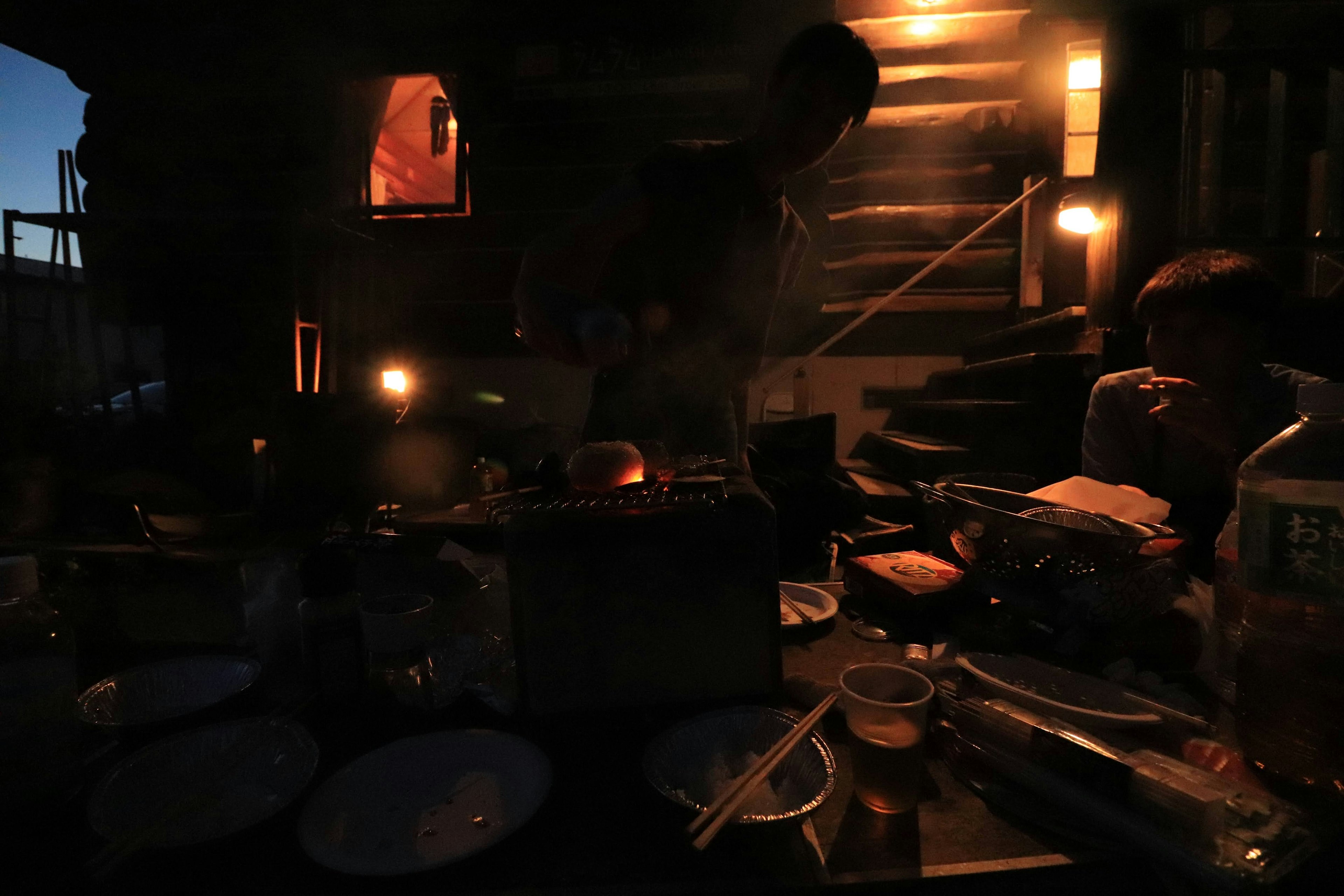 A person cooking in a dimly lit outdoor setting with scattered plates and ingredients on the table