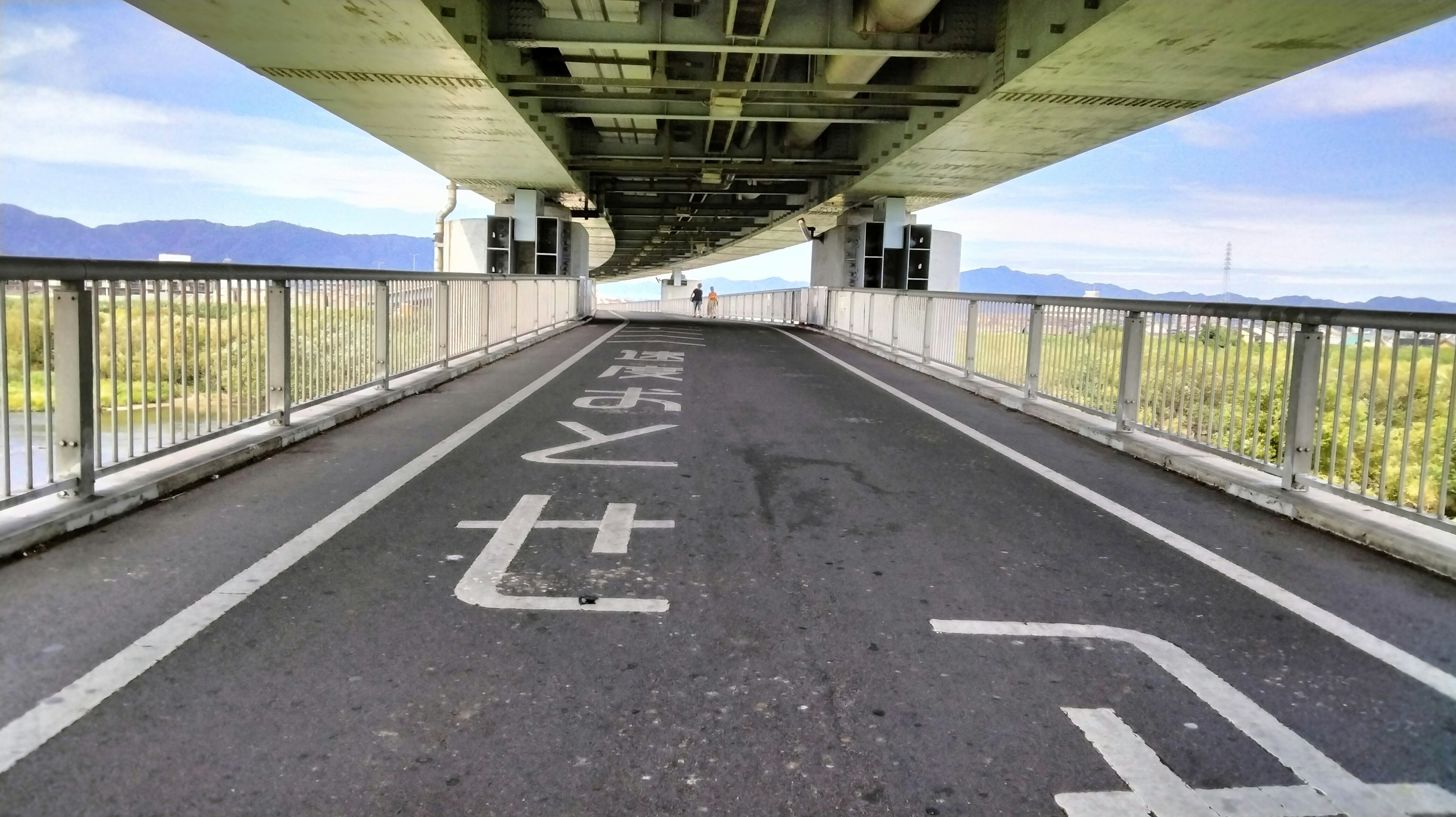 Percorso ciclabile sotto un ponte con cielo blu e montagne sullo sfondo