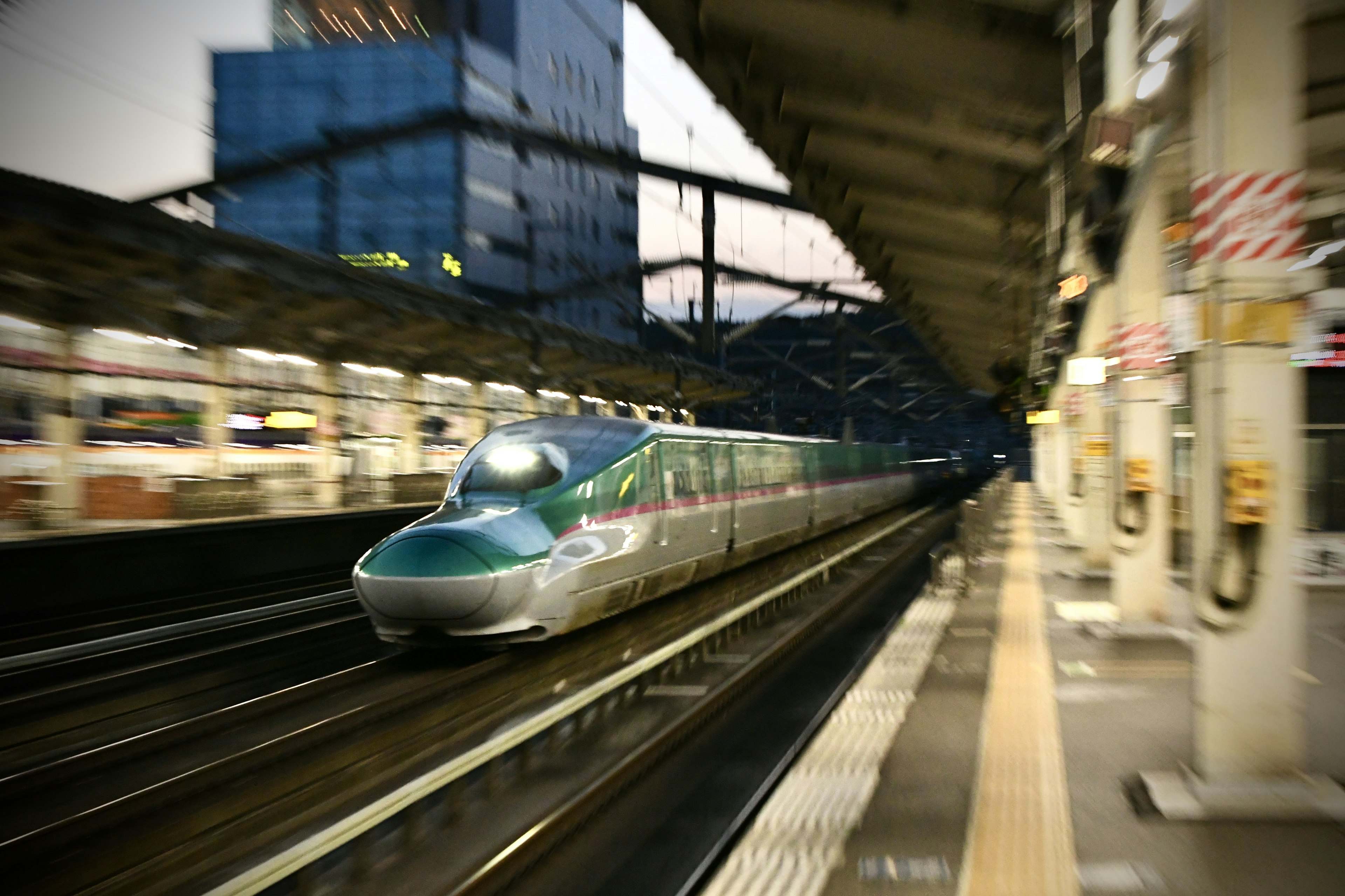 Shinkansen đi qua ga nền mờ có tòa nhà chọc trời