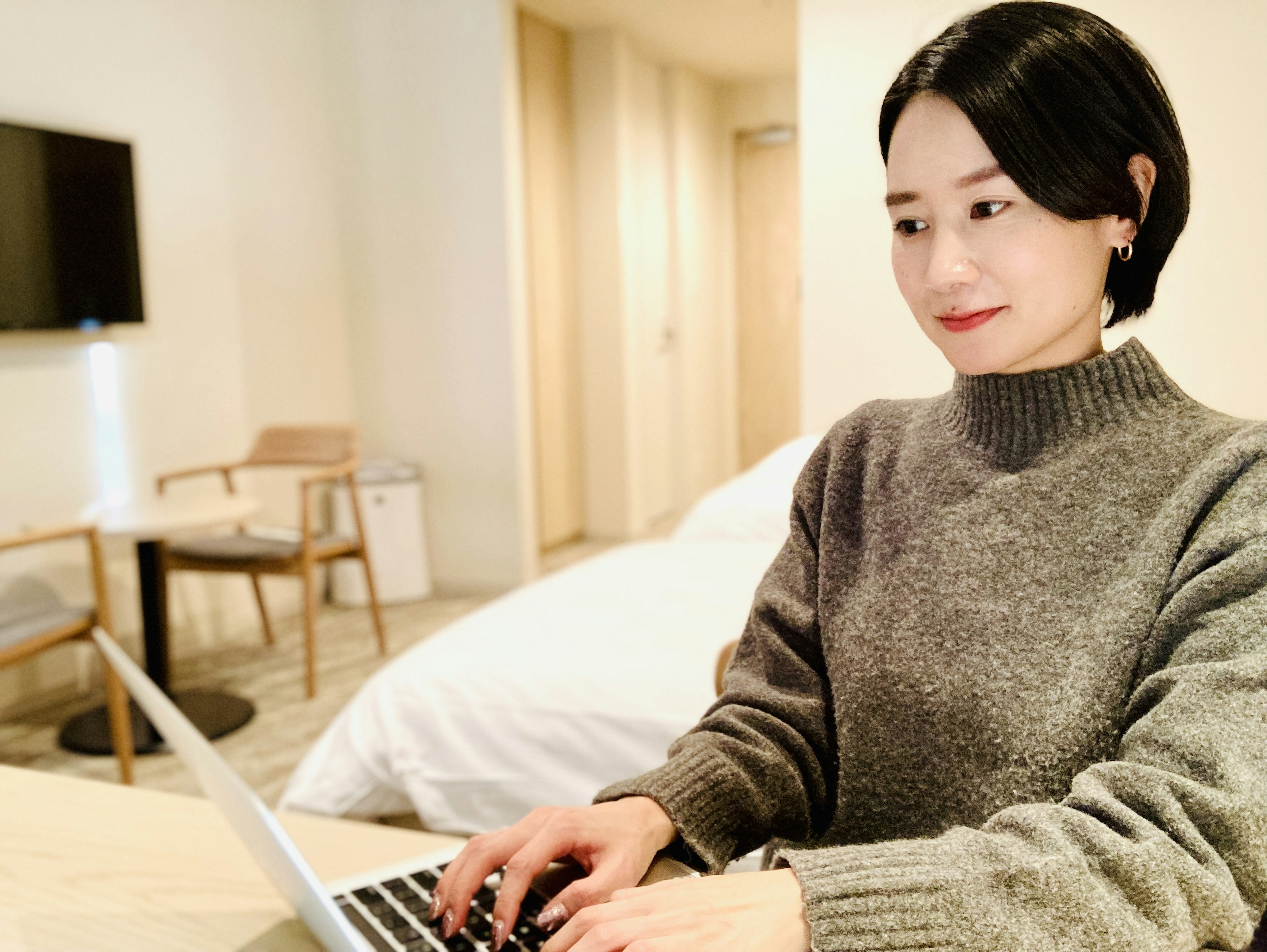 Woman working on a laptop in a hotel room wearing a gray sweater