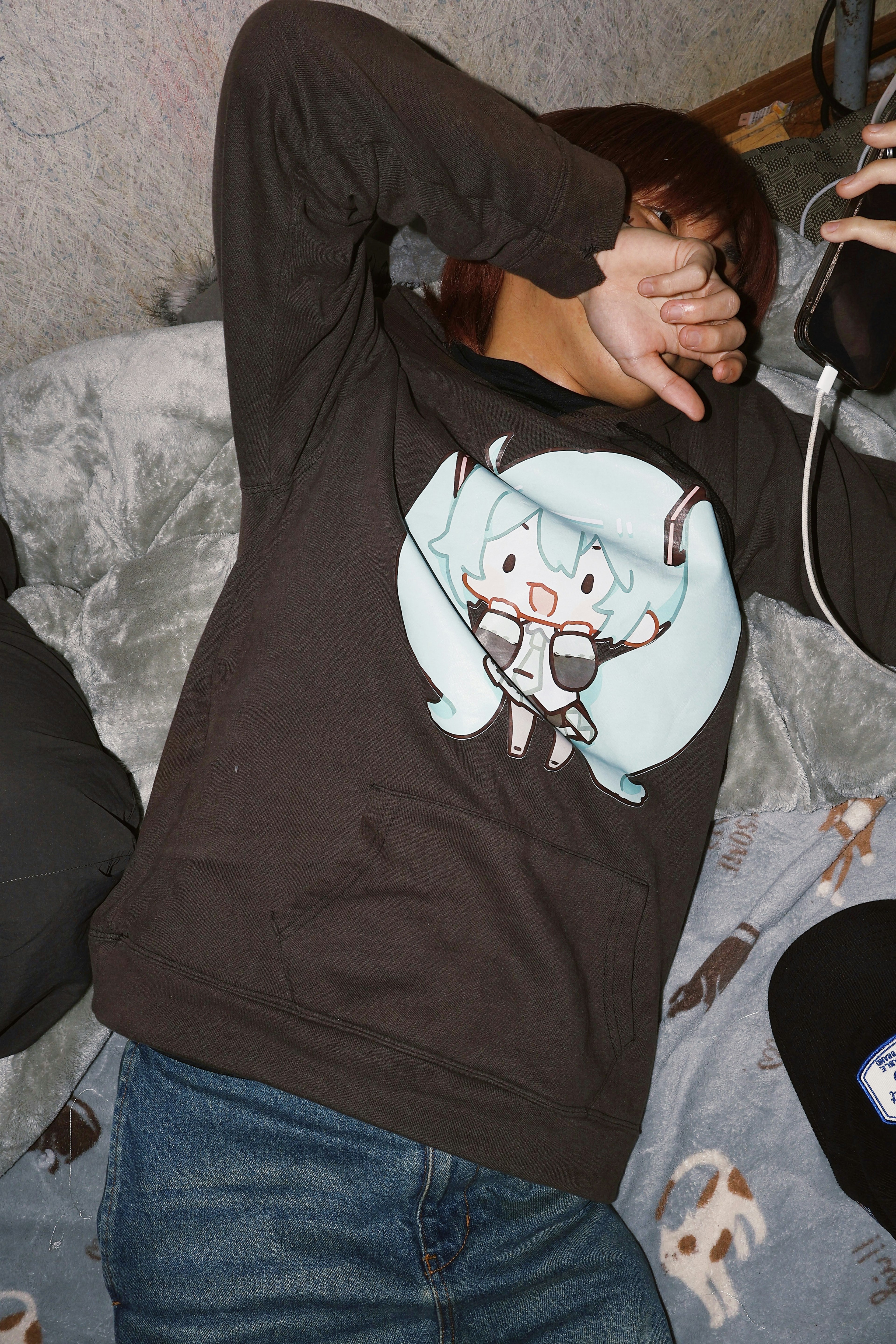 Person covering their face wearing a black long-sleeve shirt with a Hatsune Miku design