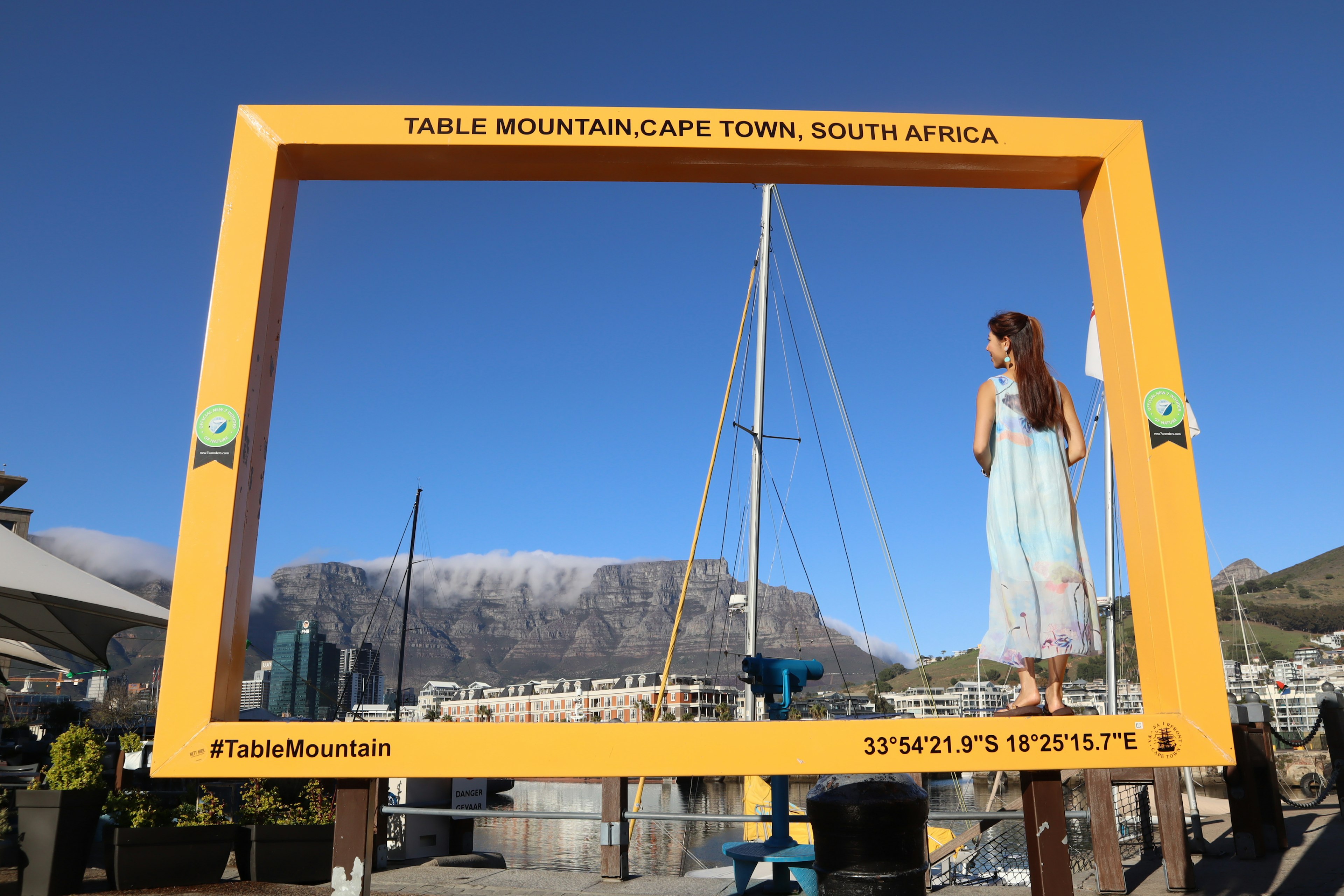 Seorang wanita berdiri di depan Gunung Meja yang dibingkai oleh border kuning di Cape Town