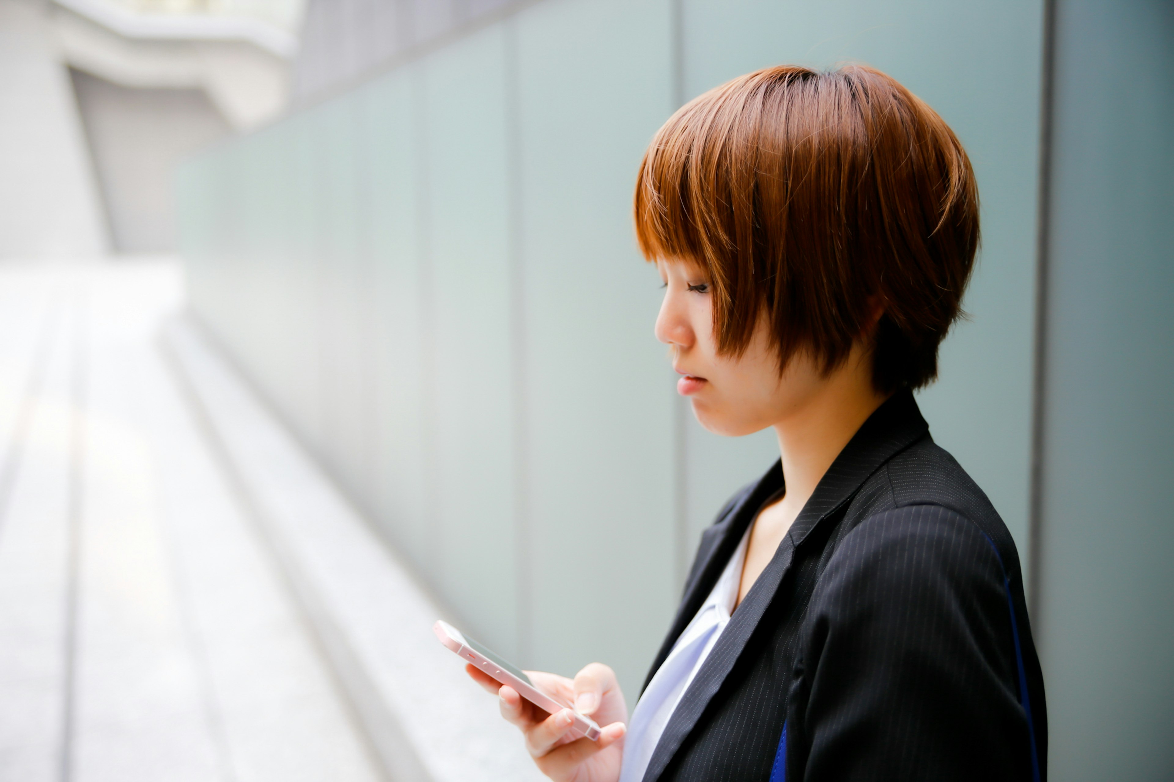 Profil wanita memegang smartphone dengan rambut cokelat pendek di latar belakang bangunan modern