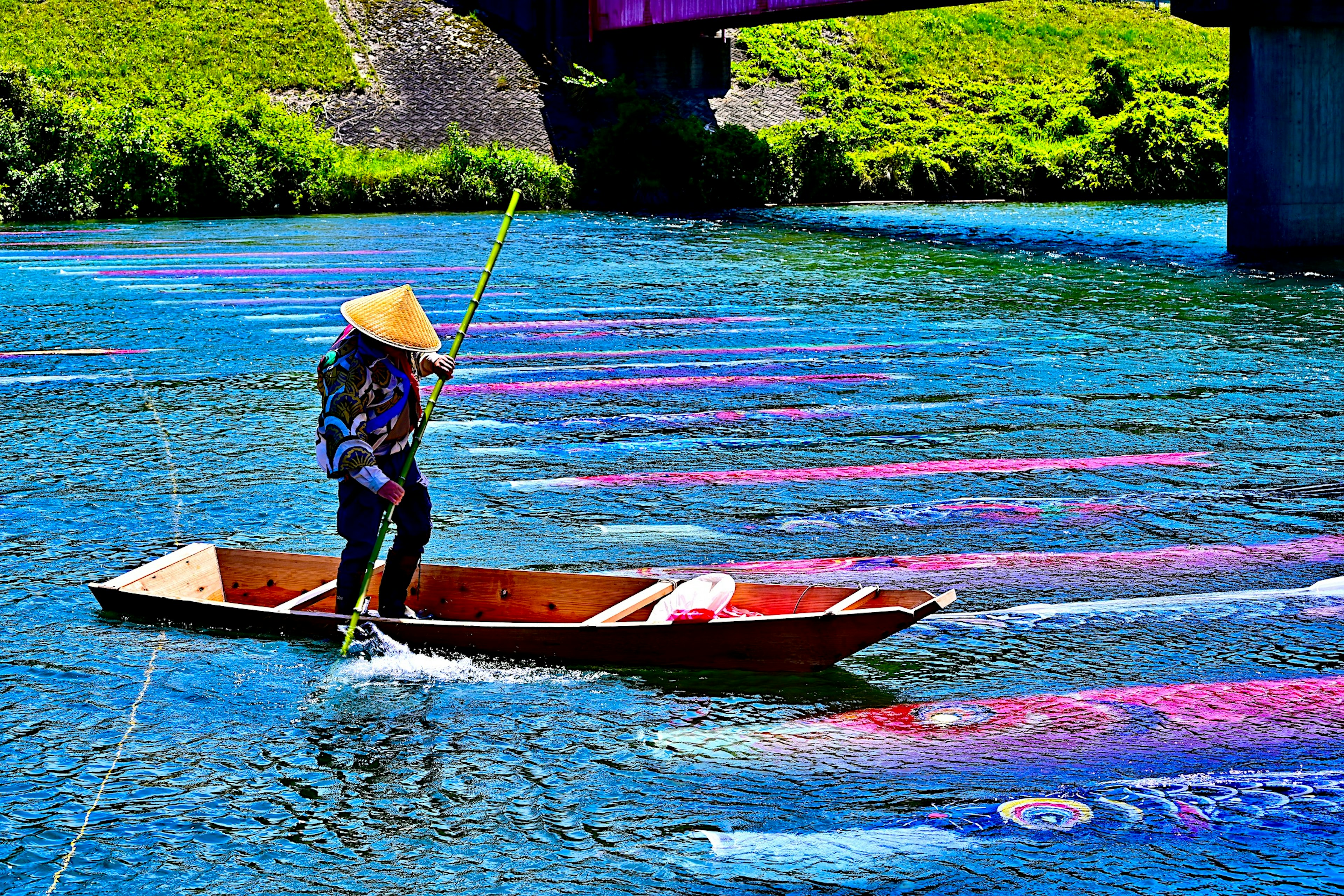 A person in a conical hat paddling a boat on a river with colorful fabrics floating