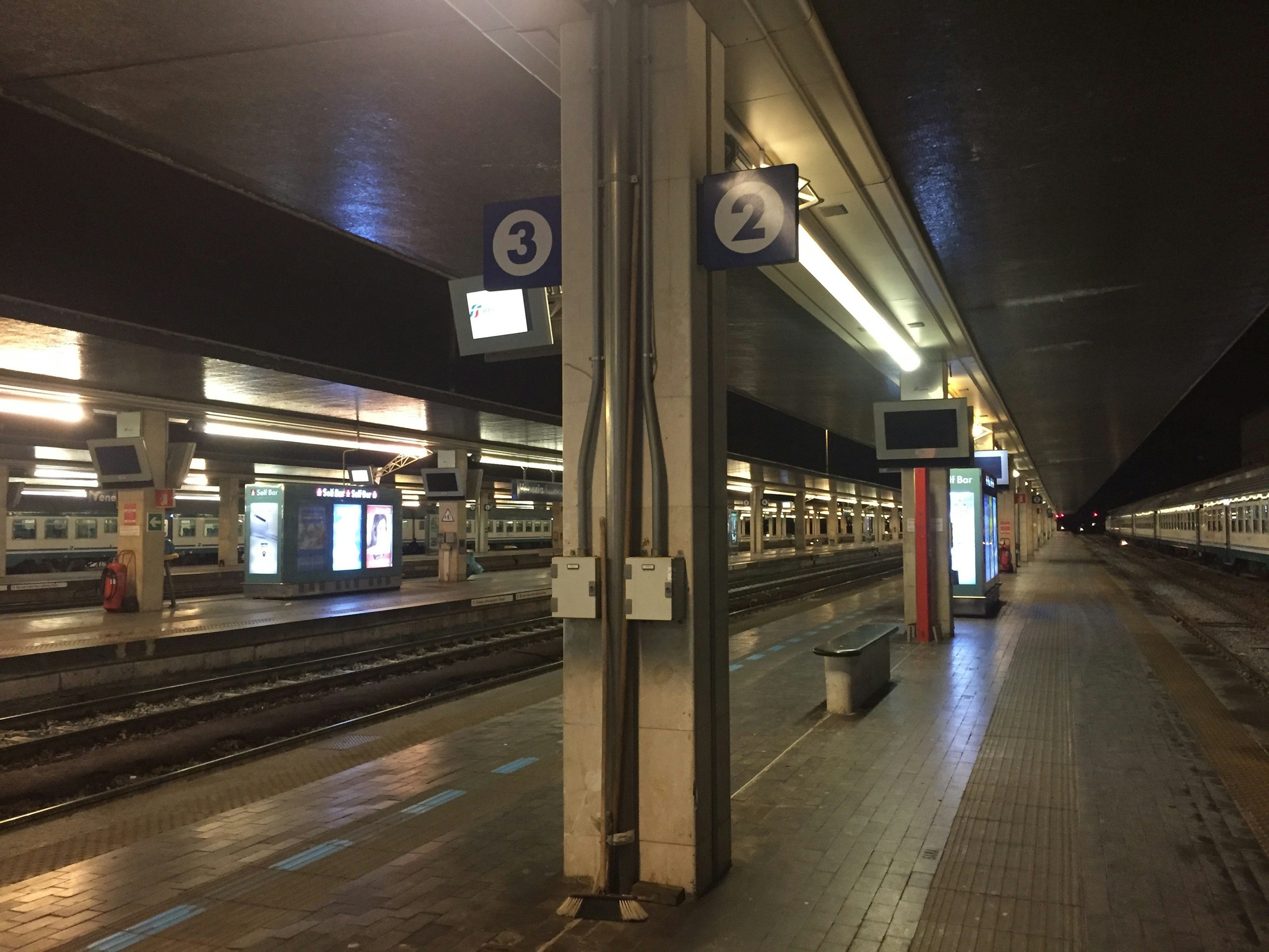 Imagen interior de una plataforma de estación de tren con vías y pantallas de información