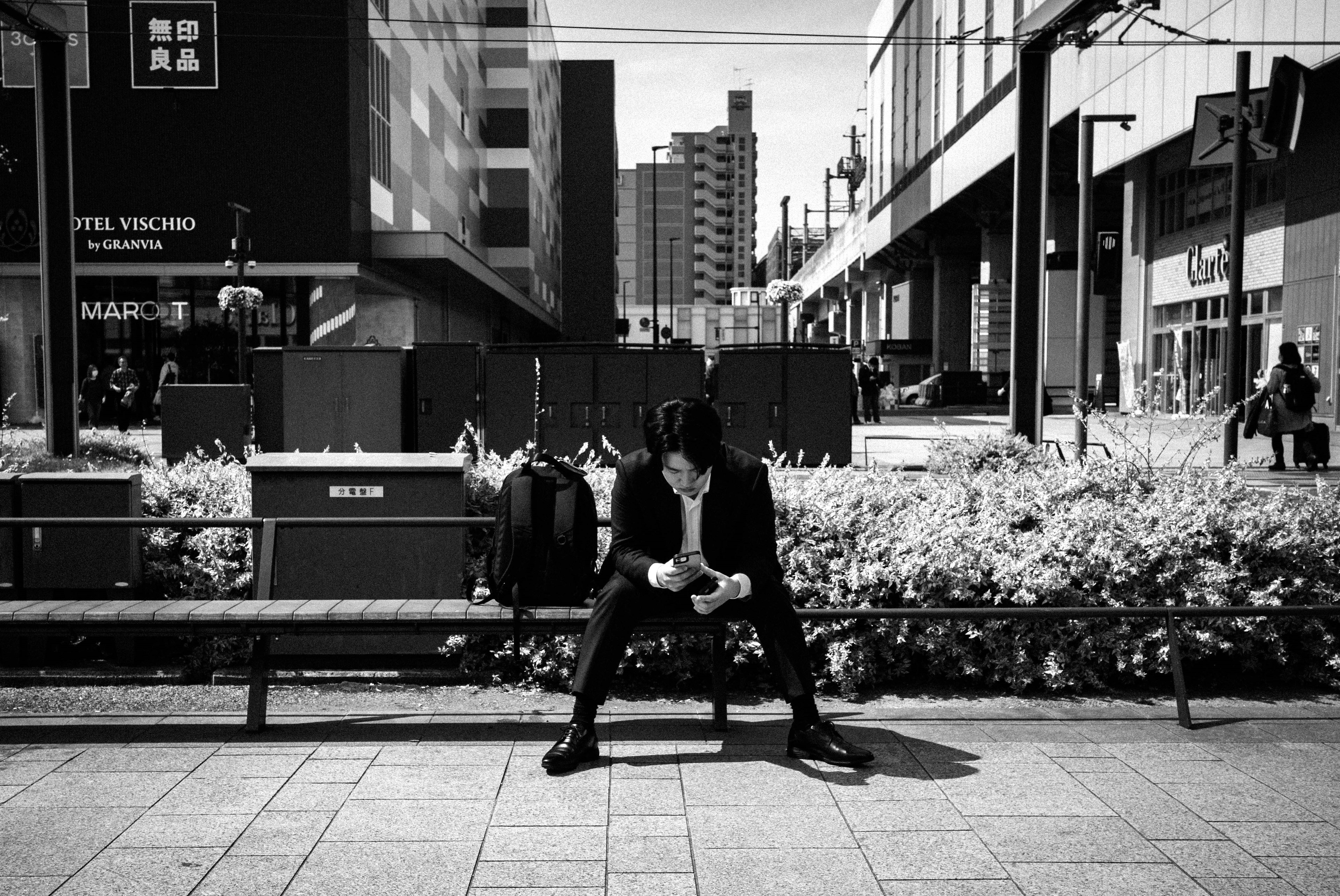 Un hombre sentado en un banco en una ciudad mirando su teléfono inteligente en blanco y negro