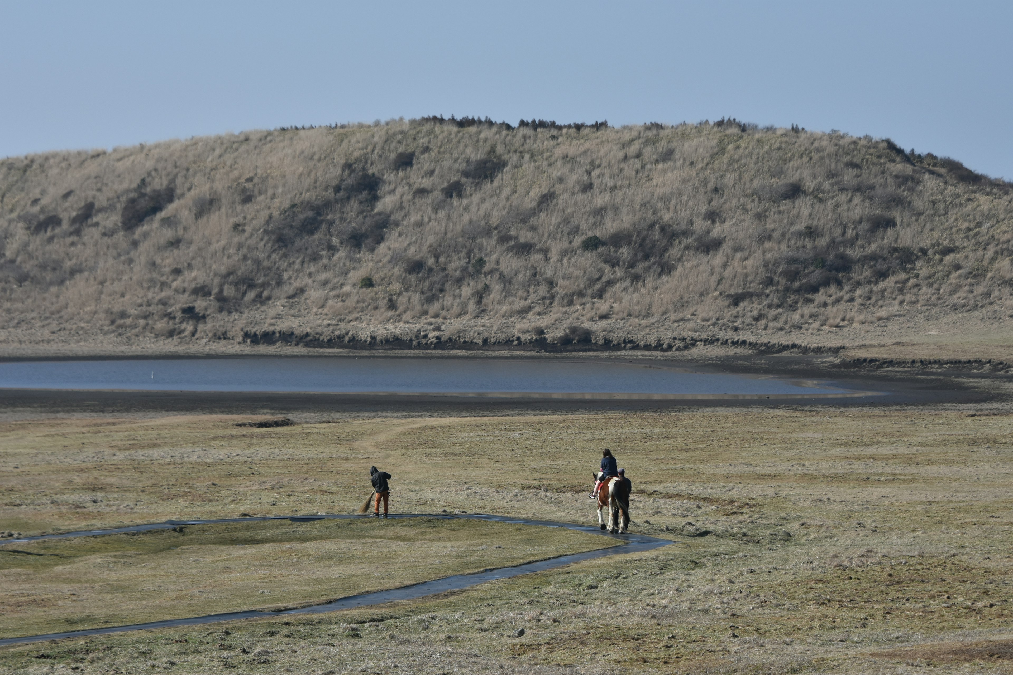 People walking in a grassy landscape with a small hill and calm lake in the background