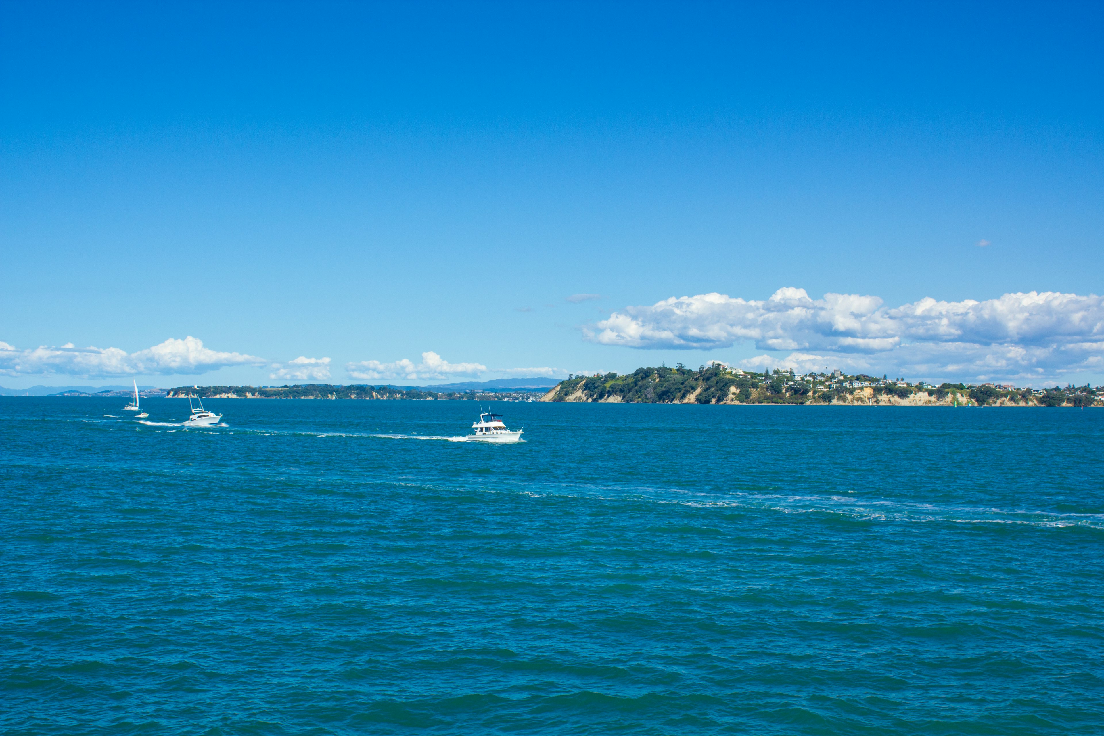 Boats cruising on blue water under a clear sky with white clouds and an island in the distance